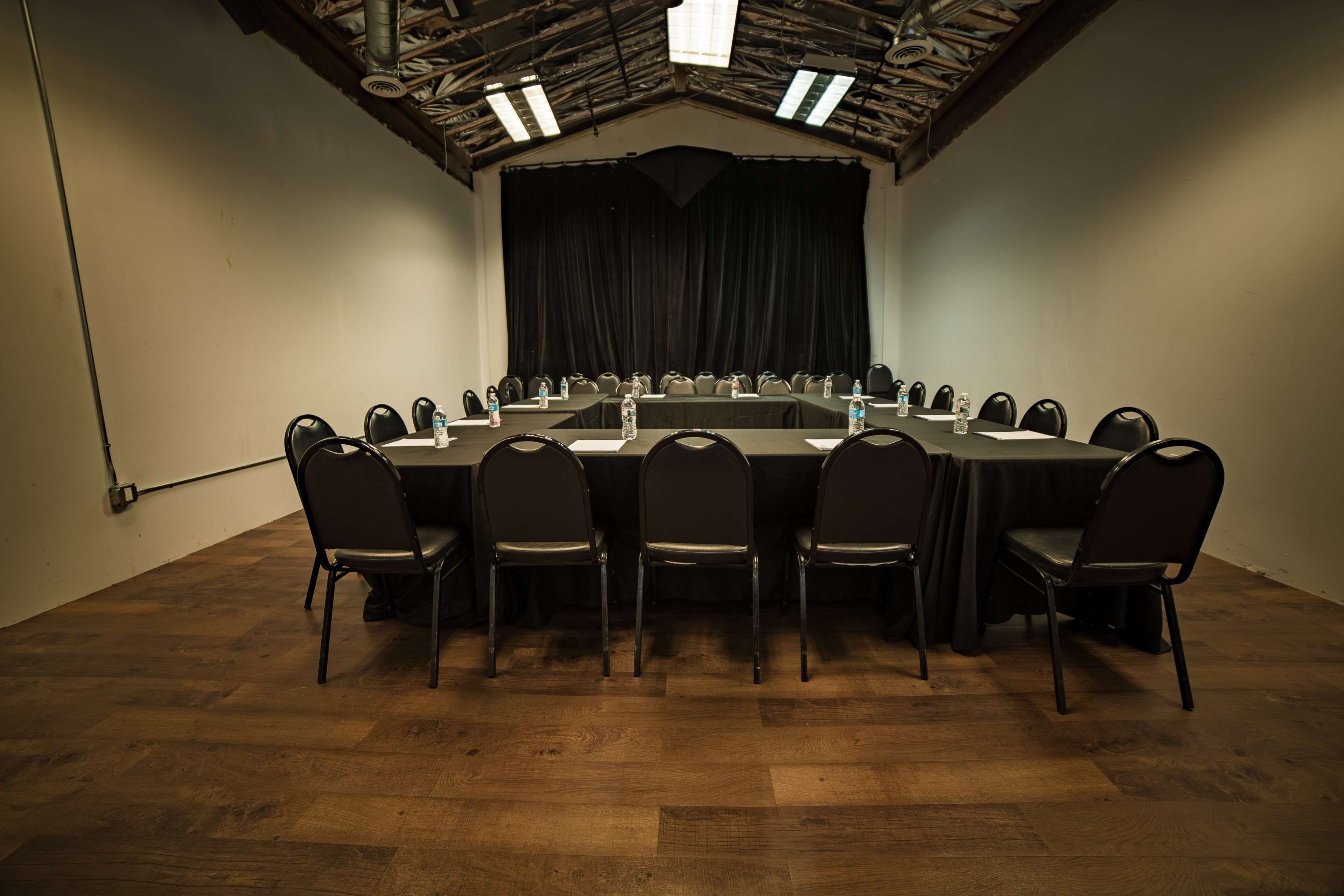 A large, rectangular table is set up in a room with wooden floors, surrounded by black chairs and draped in black cloth, facing a black curtain backdrop.