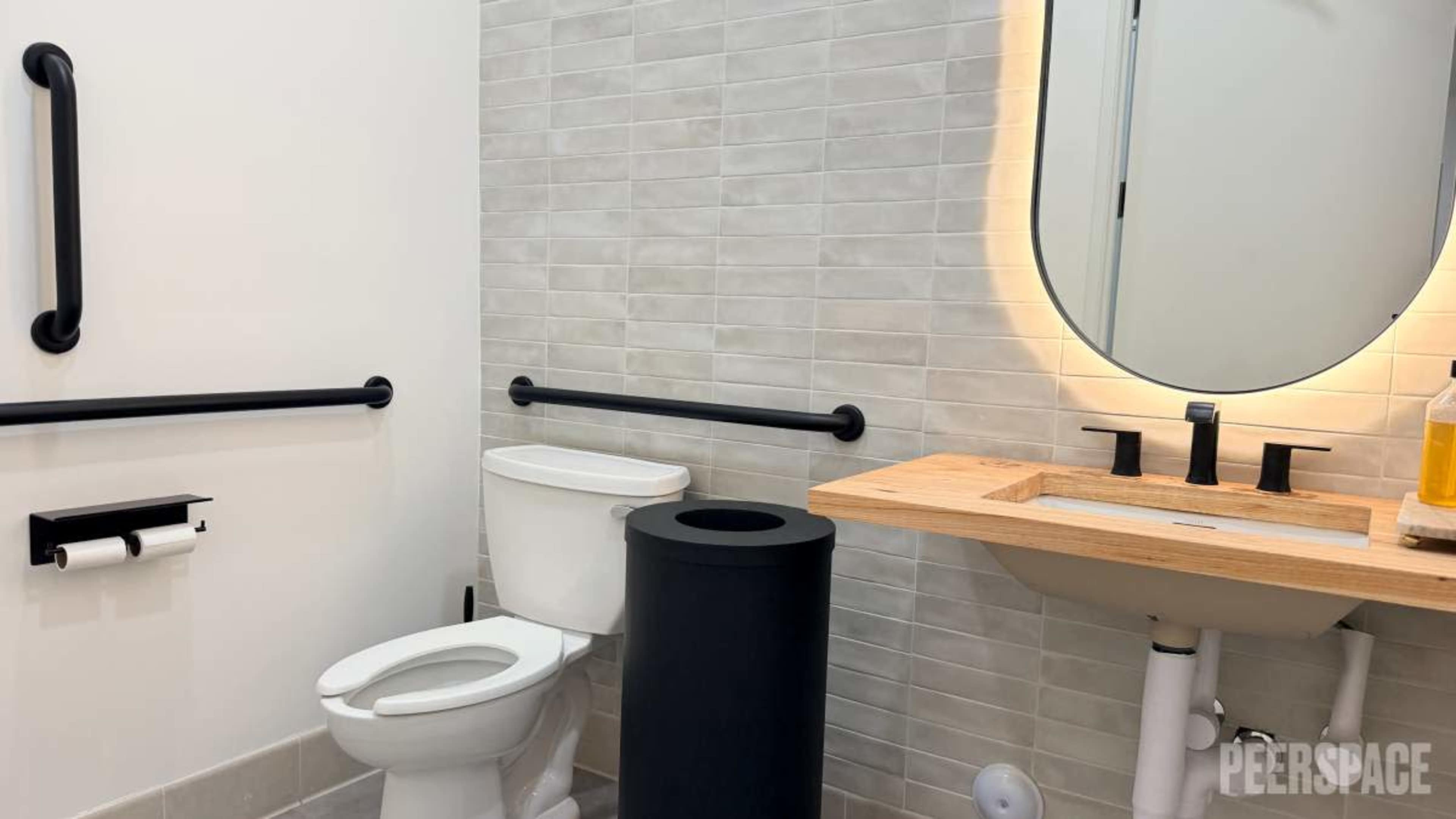 The image shows a modern bathroom featuring a toilet, a sink with a wooden countertop, and a large round mirror against tiled walls.