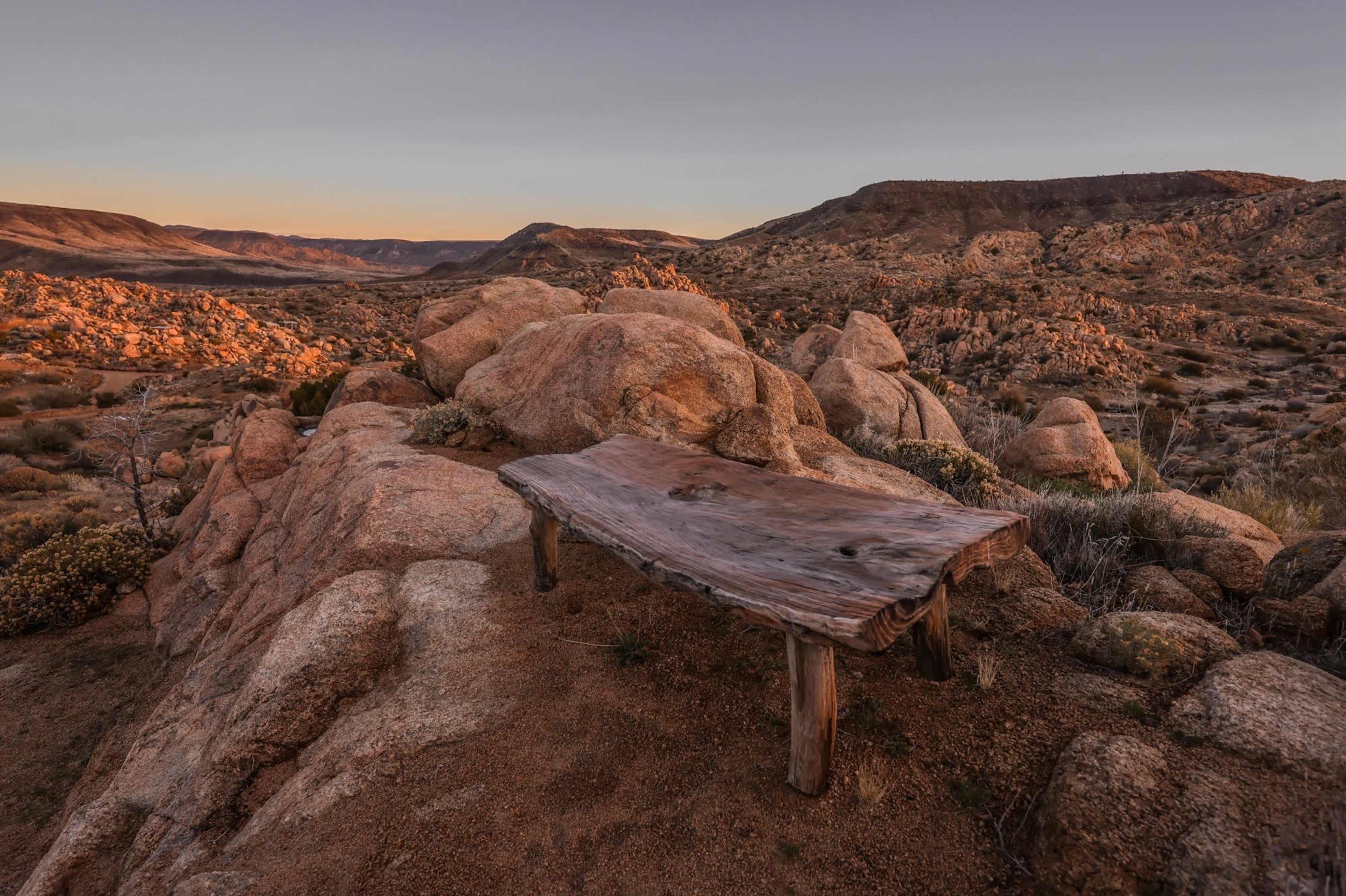 A wooden bench is positioned on a rocky outcrop overlooking a desert landscape at sunset.