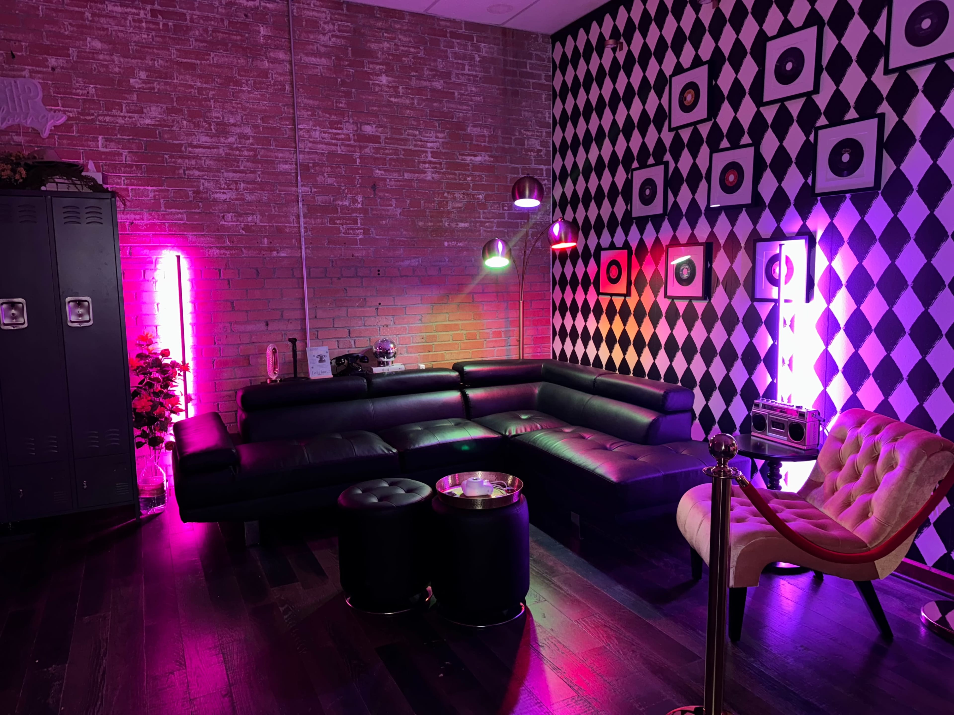 The image shows a modern lounge area featuring a black sectional sofa, round black tables, and a stylish armchair, with a brick wall and a checkered wall design illuminated by colorful lights.