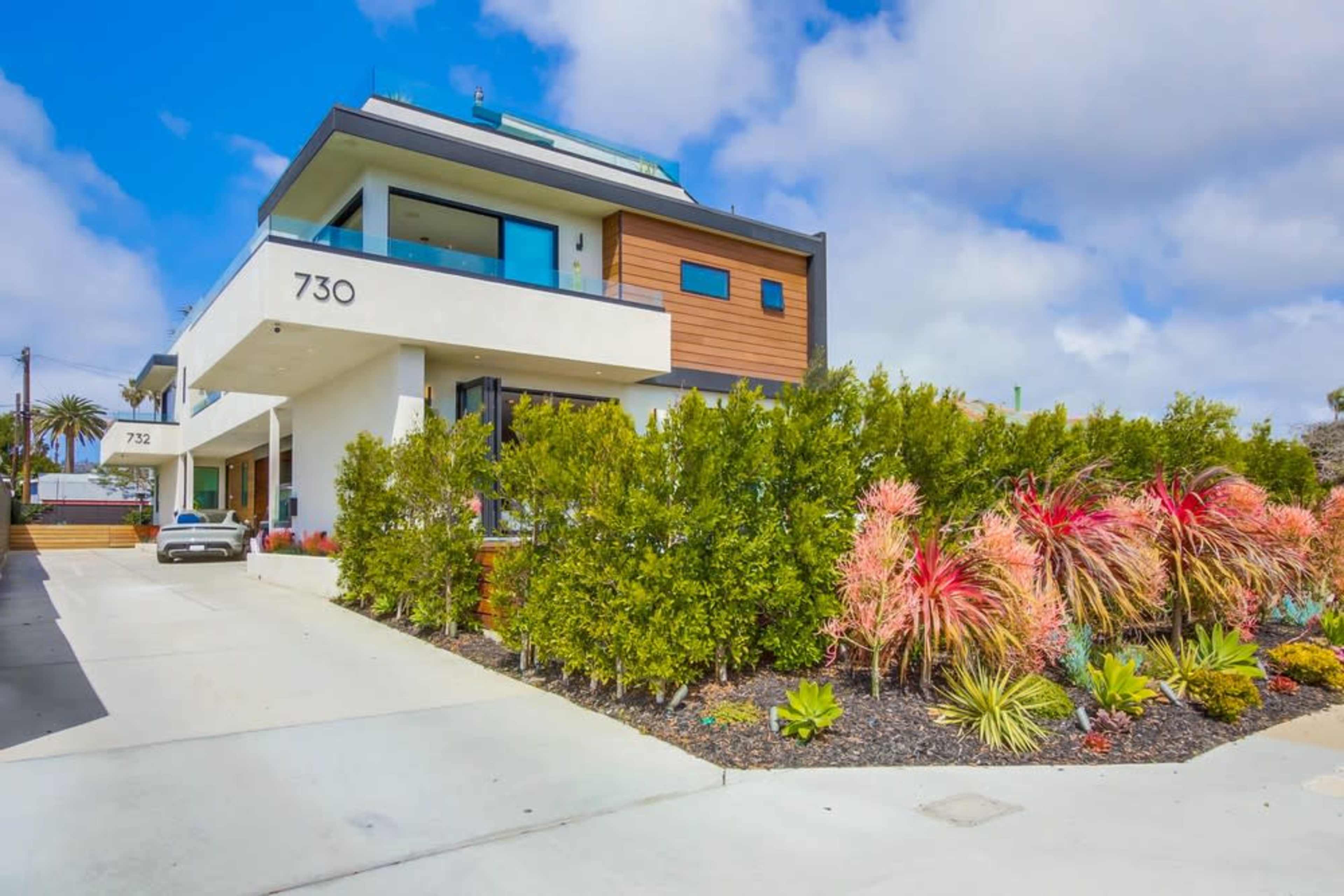 A modern two-story house with a landscaped front garden featuring various plants and a driveway leading to the entrance.