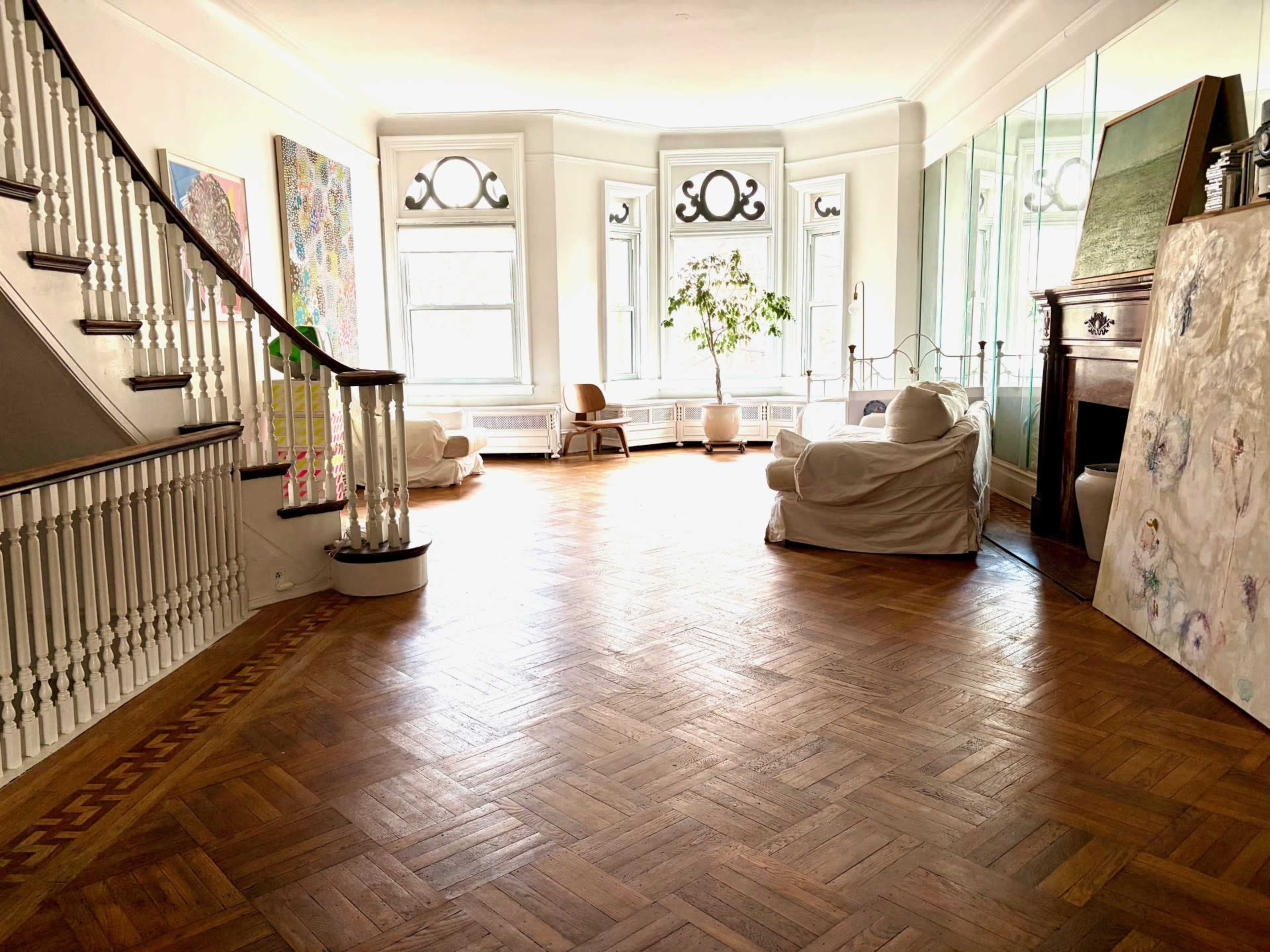 The image shows a spacious room featuring wooden flooring, large windows, a staircase, and various pieces of artwork on the walls.