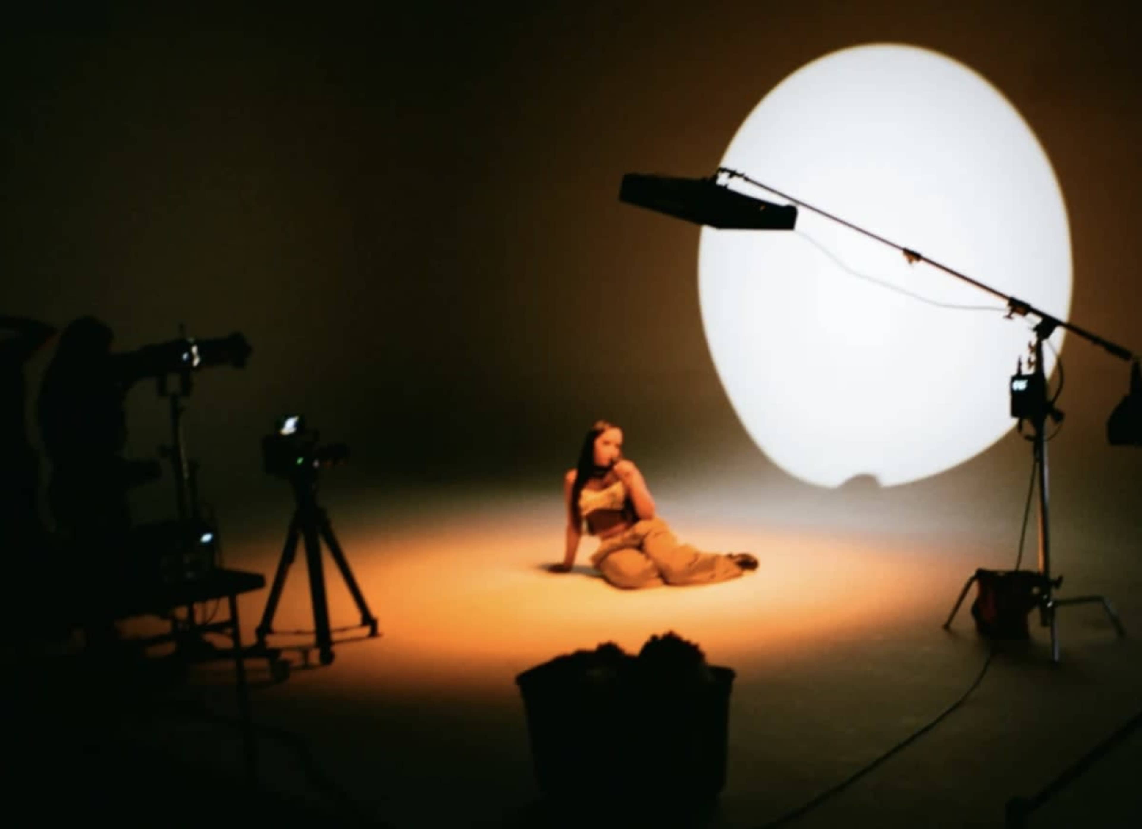 A model poses on a studio floor while illuminated by a large circular light, with cameras and lighting equipment positioned around her.