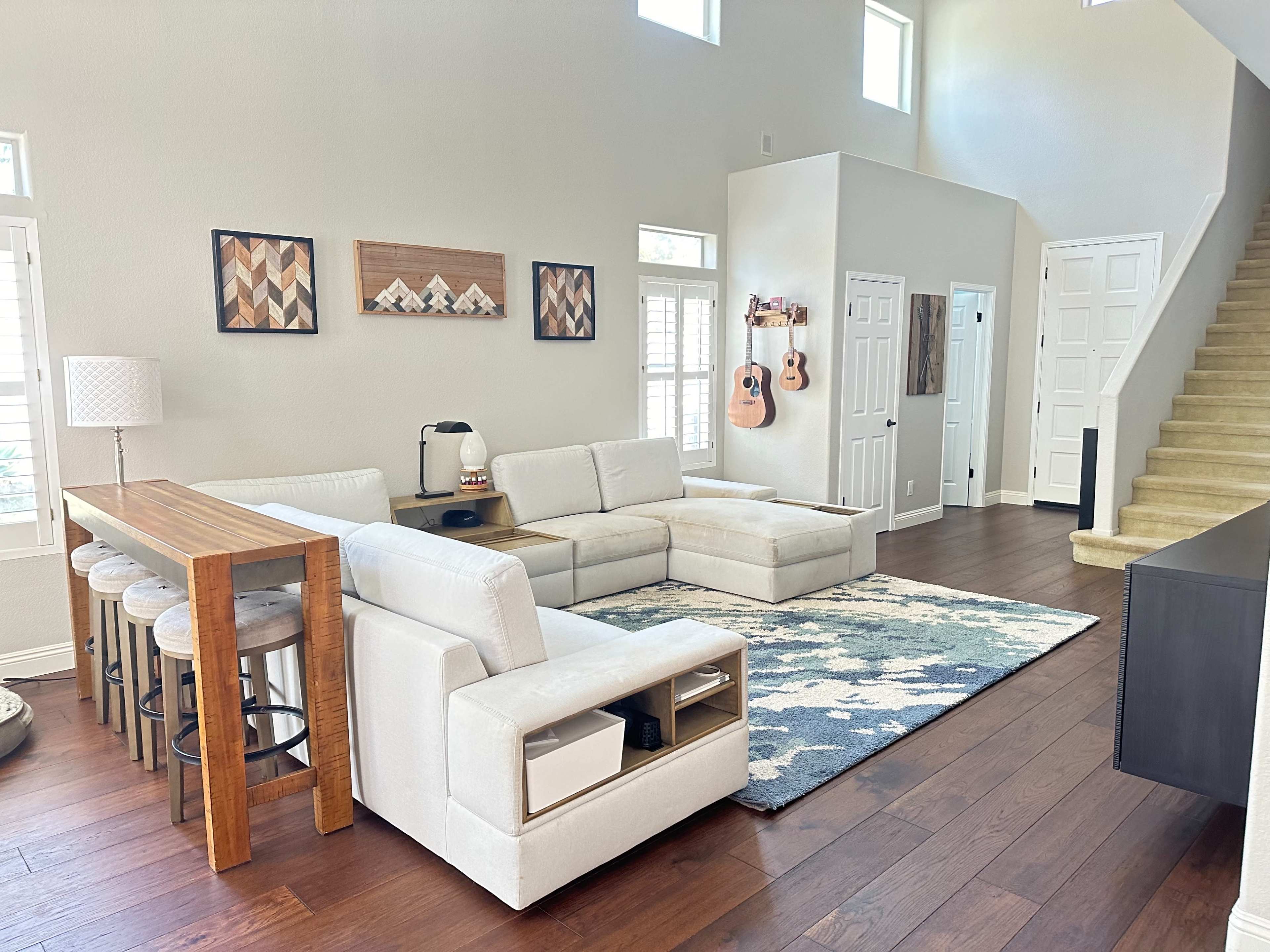 The spacious living room features a sectional sofa, a wooden dining table with stools, a decorative rug, and a staircase leading to an upper level.
