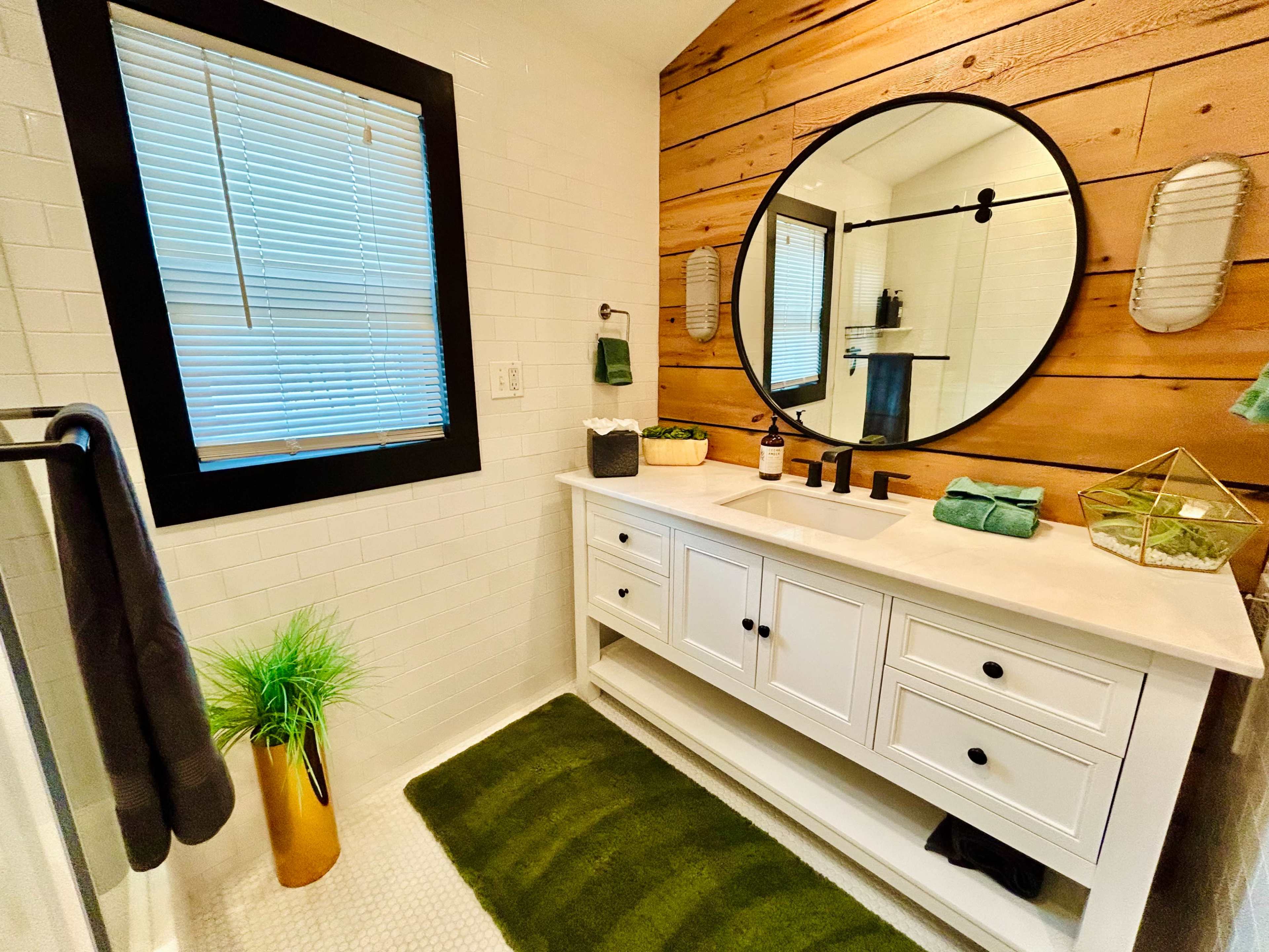 The image shows a modern bathroom with a wooden accent wall, a circular mirror above a white vanity, and green decorative elements.