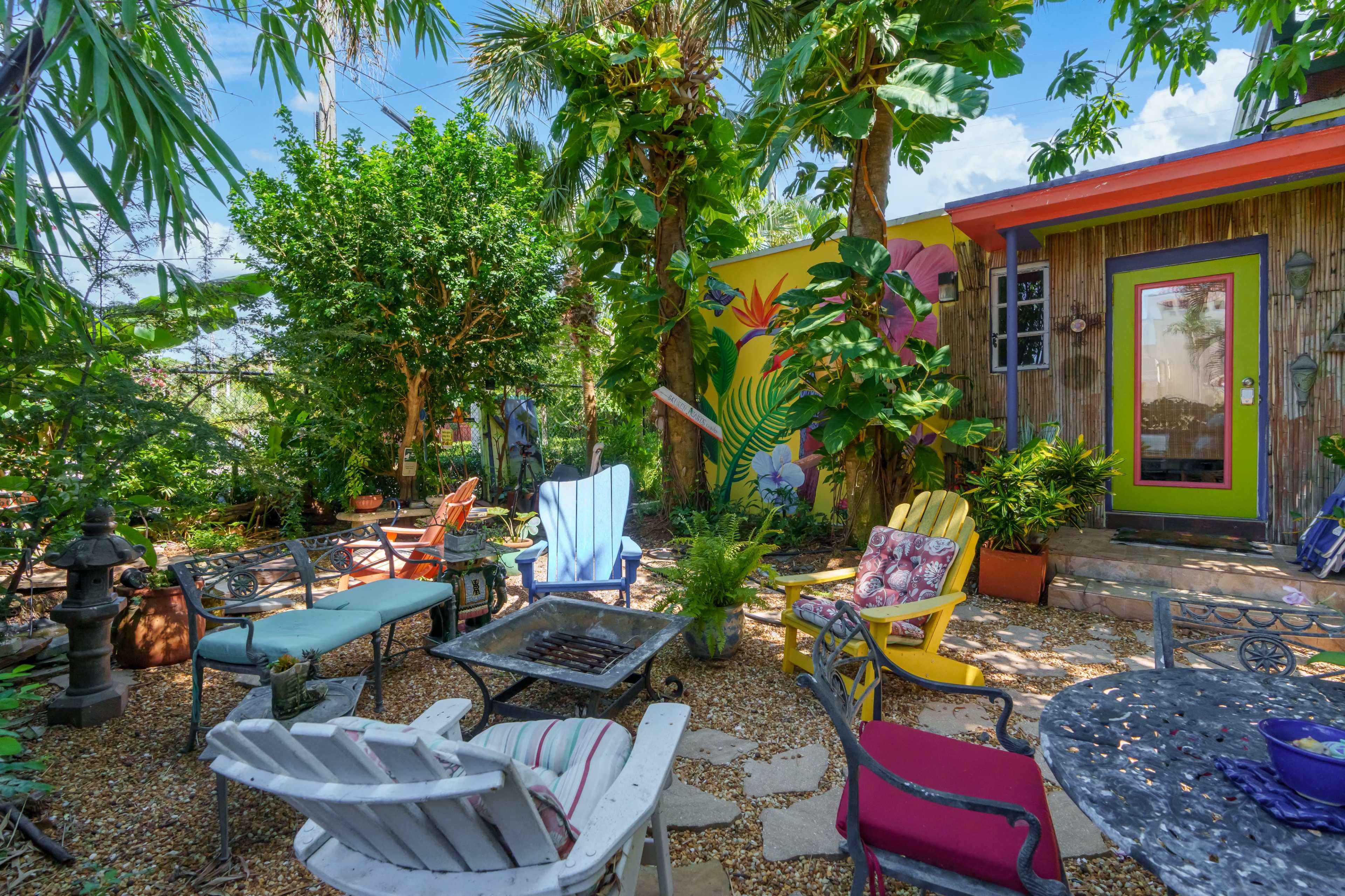 A vibrant garden setting with various colorful chairs arranged around a fire pit, surrounded by lush greenery and a mural on a nearby wall.