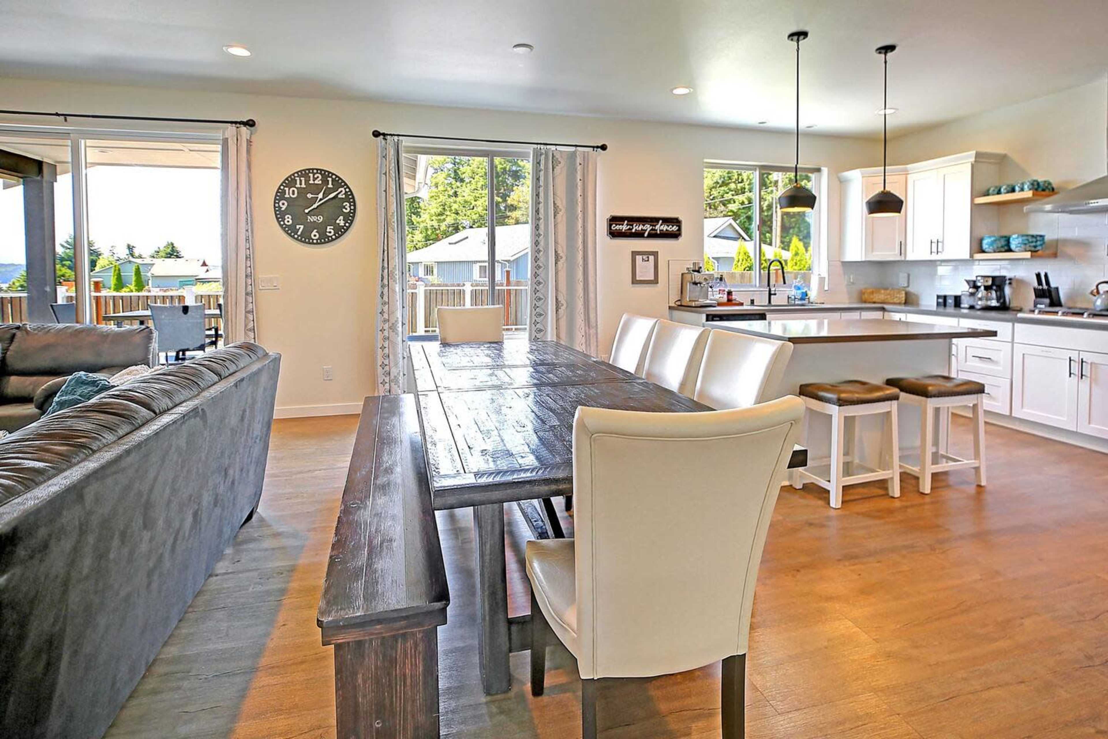 The image shows a modern open-concept living and dining area featuring a wooden dining table, upholstered chairs, and a well-equipped kitchen in the background.