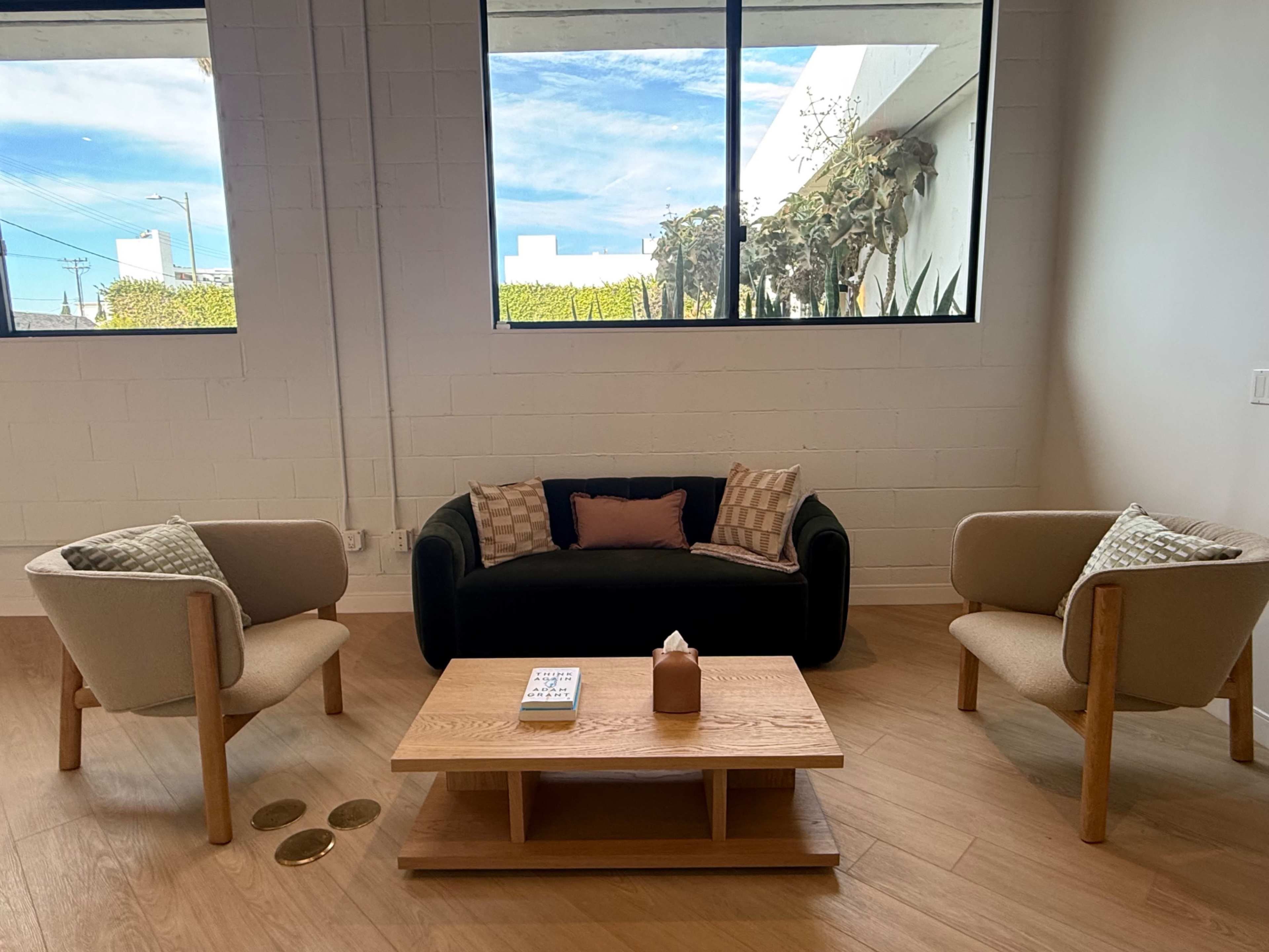 A modern living space features a black sofa with decorative cushions, two beige armchairs, and a wooden coffee table, all set against a backdrop of large windows letting in natural light.