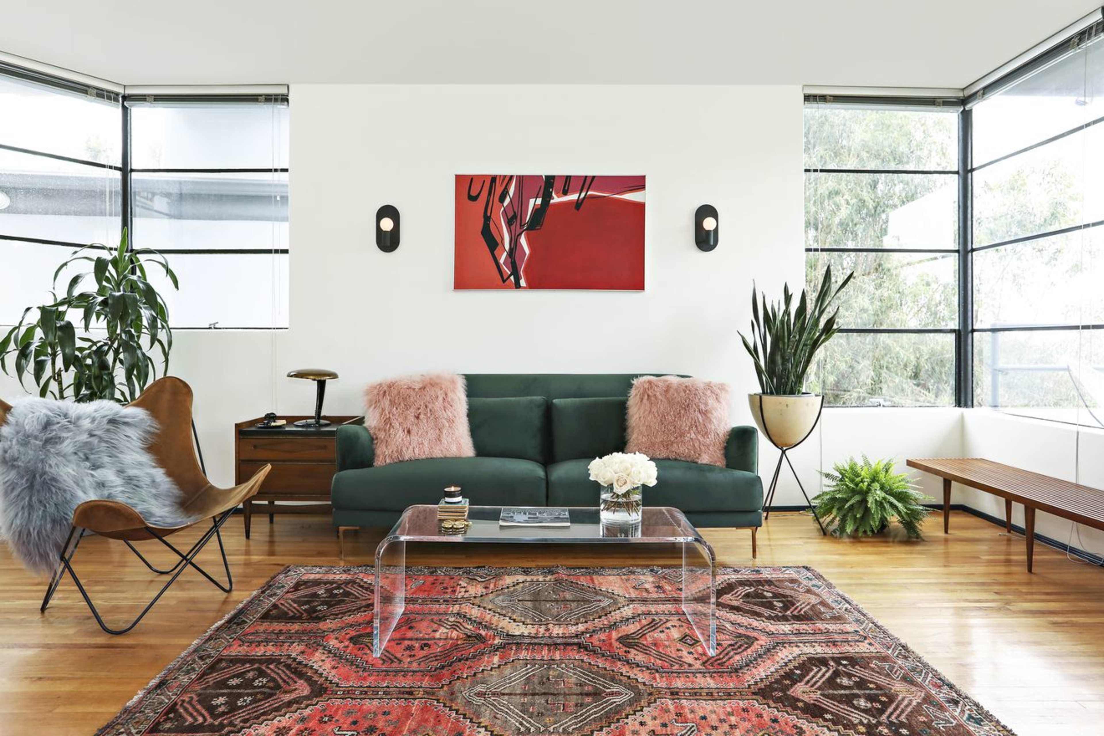 A modern living room features a green sofa with pink cushions, a glass coffee table, an abstract painting on the wall, and various plants in the corners.