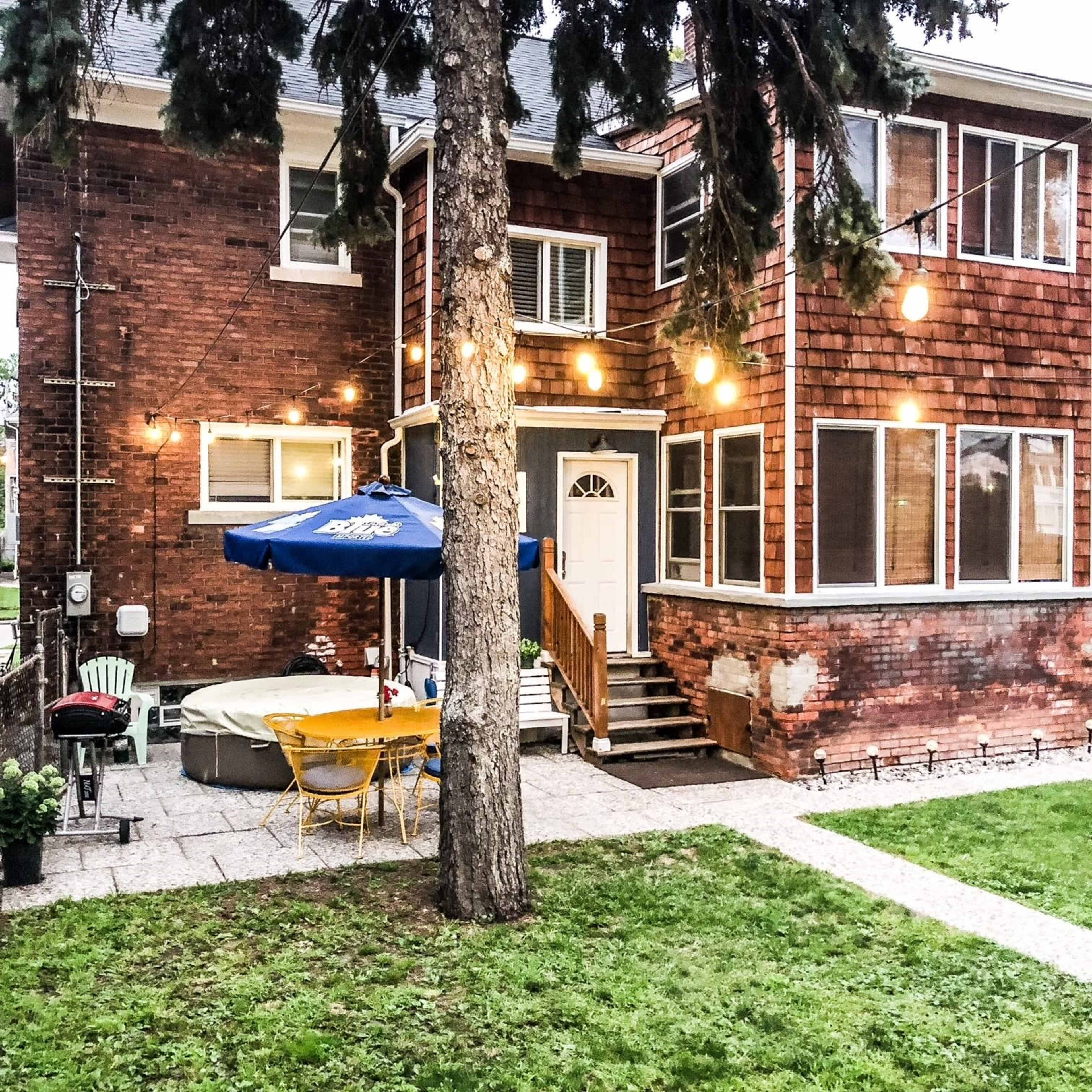 The back of a two-story brick house with a patio area featuring a hot tub and outdoor dining set under string lights.