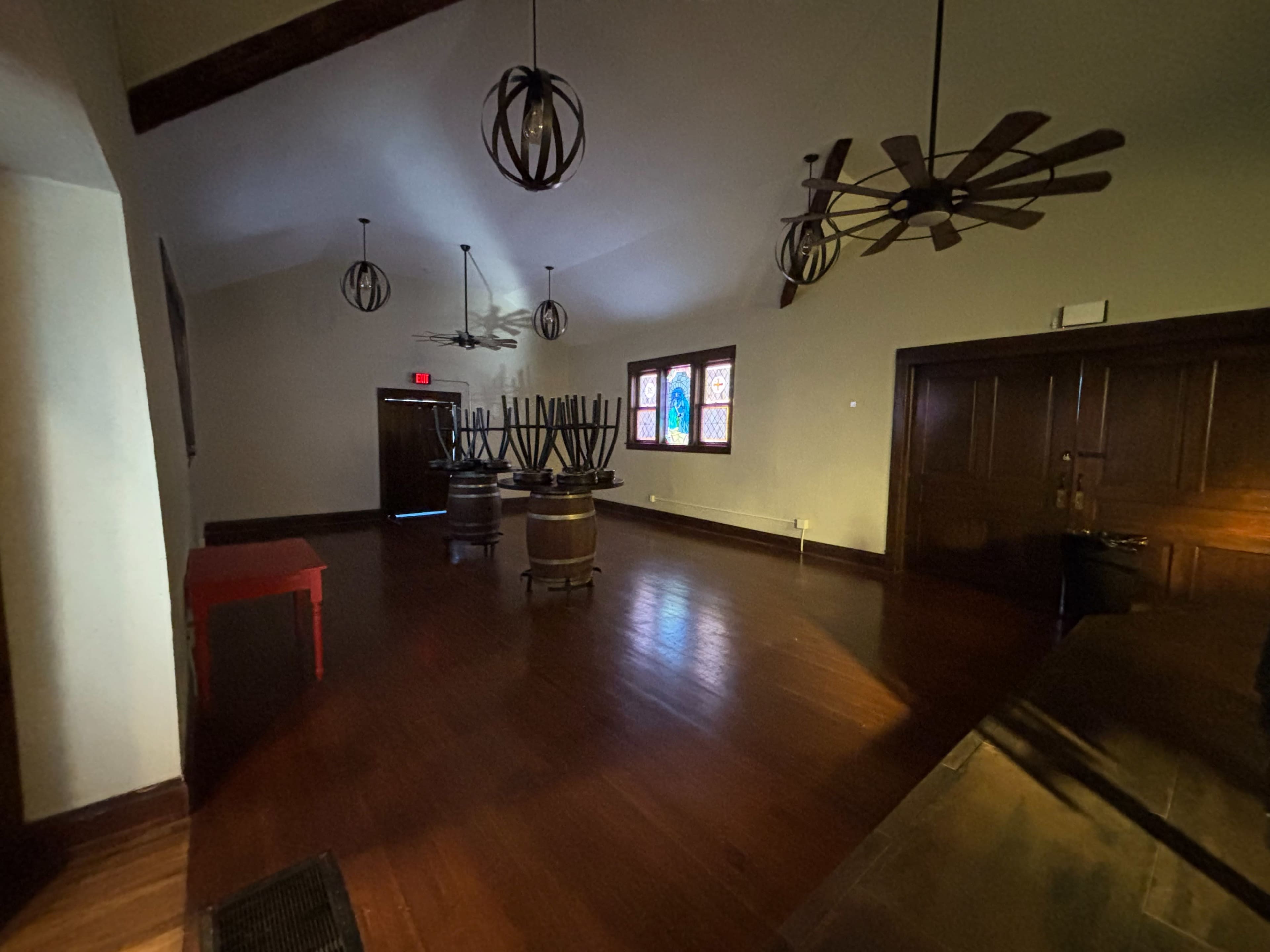 The image shows a dimly lit interior room featuring wooden floors, a few tall tables, and stained glass windows.