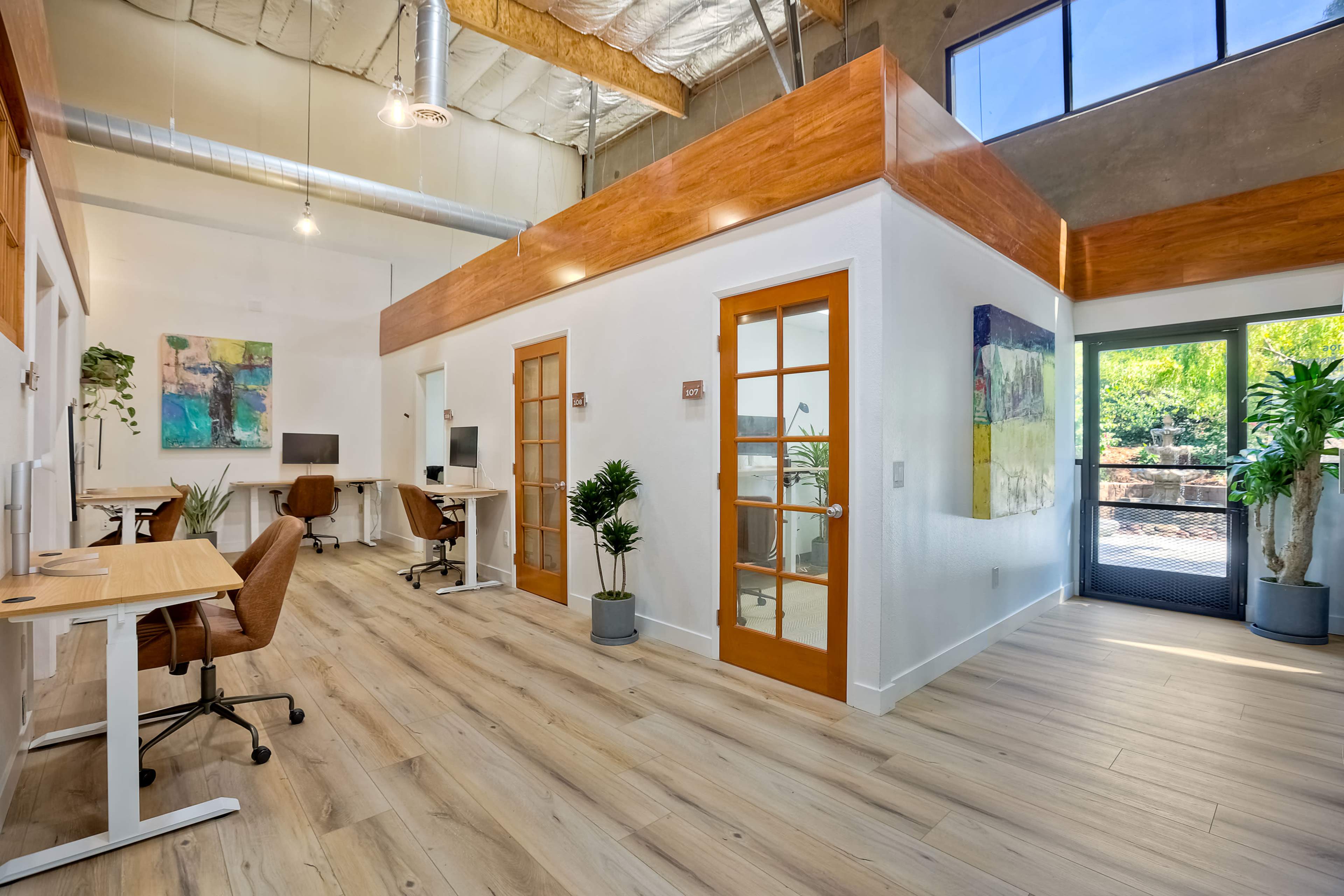 The image shows a modern office space with wooden accents, large windows, and separate workstations.