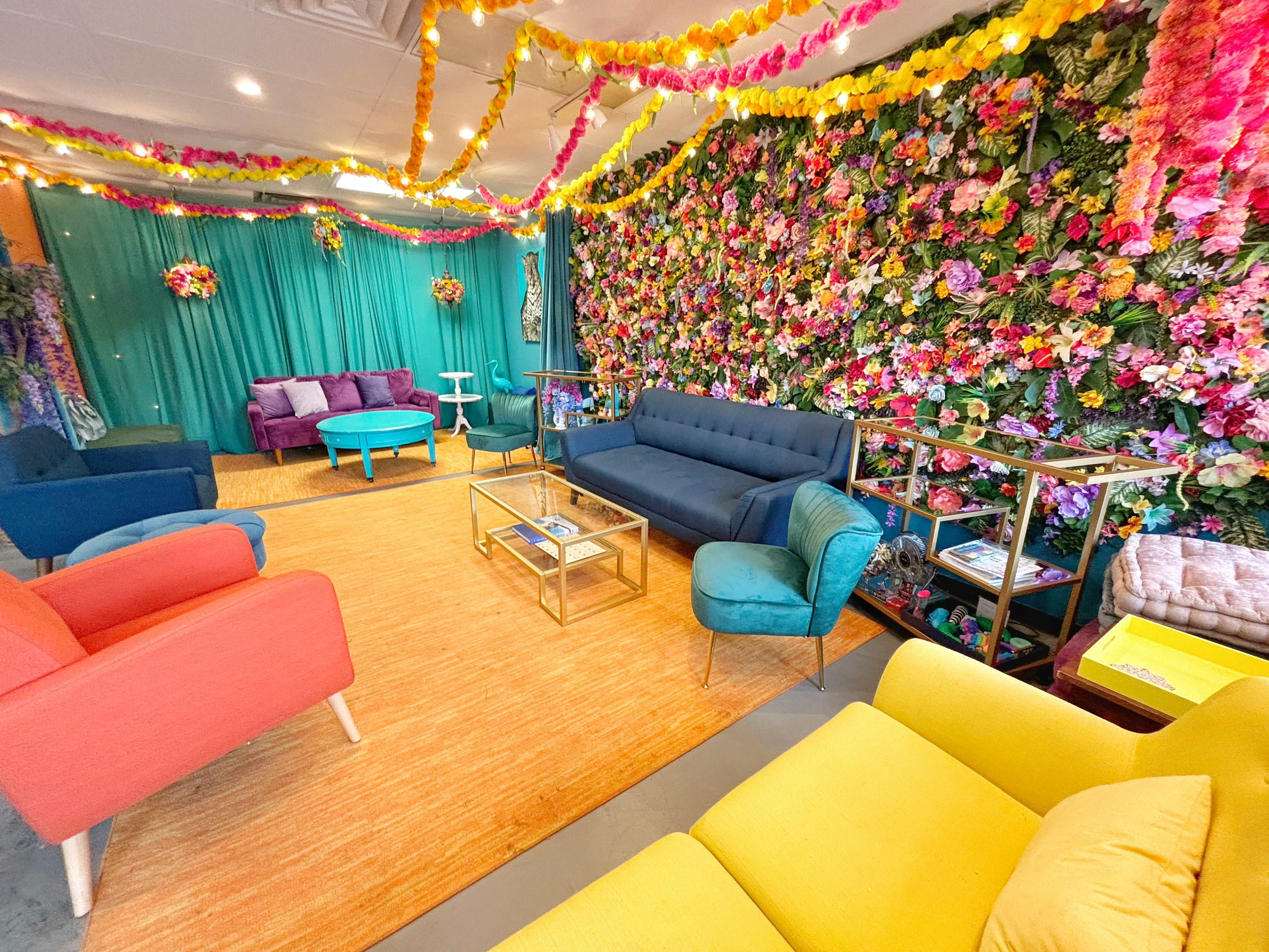 The image depicts a colorful lounge area adorned with a flower-covered wall, various seating options in bright hues, and decorative lighting.