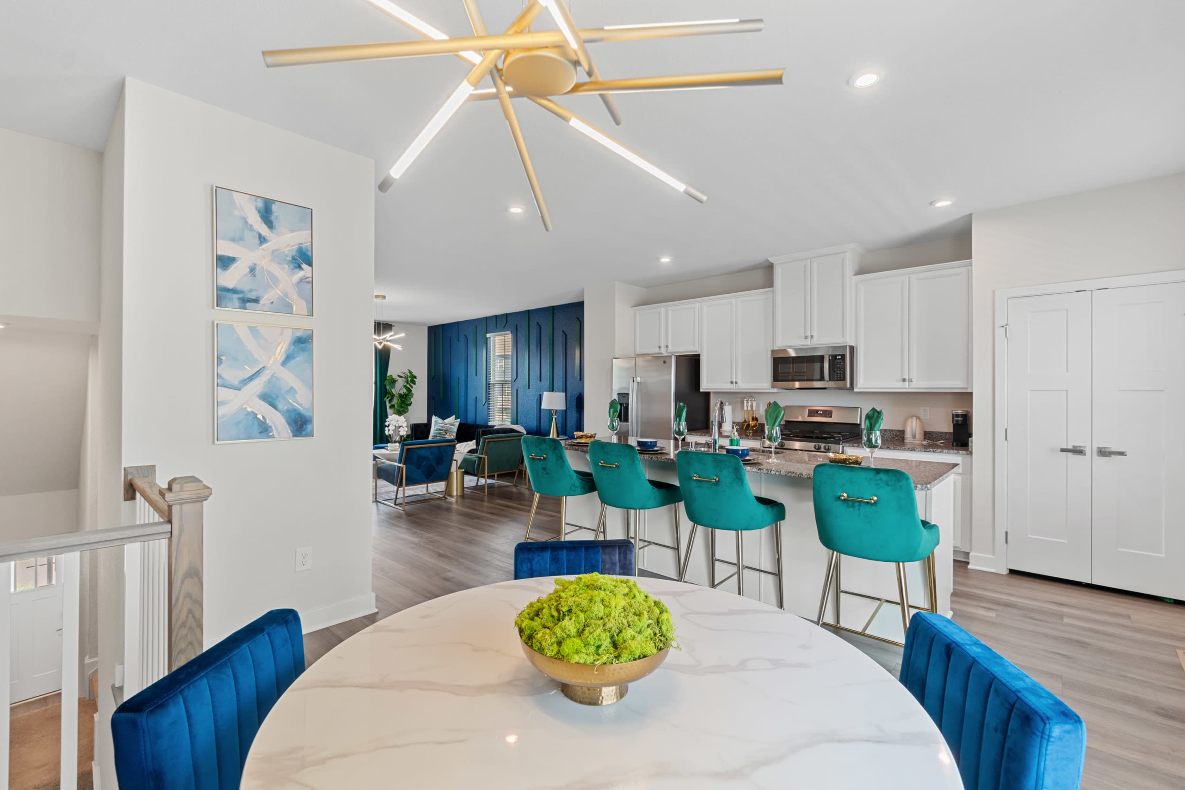 A modern open-concept living space with a dining area featuring a round marble table and green chairs, leading into a kitchen with white cabinetry and stainless steel appliances.