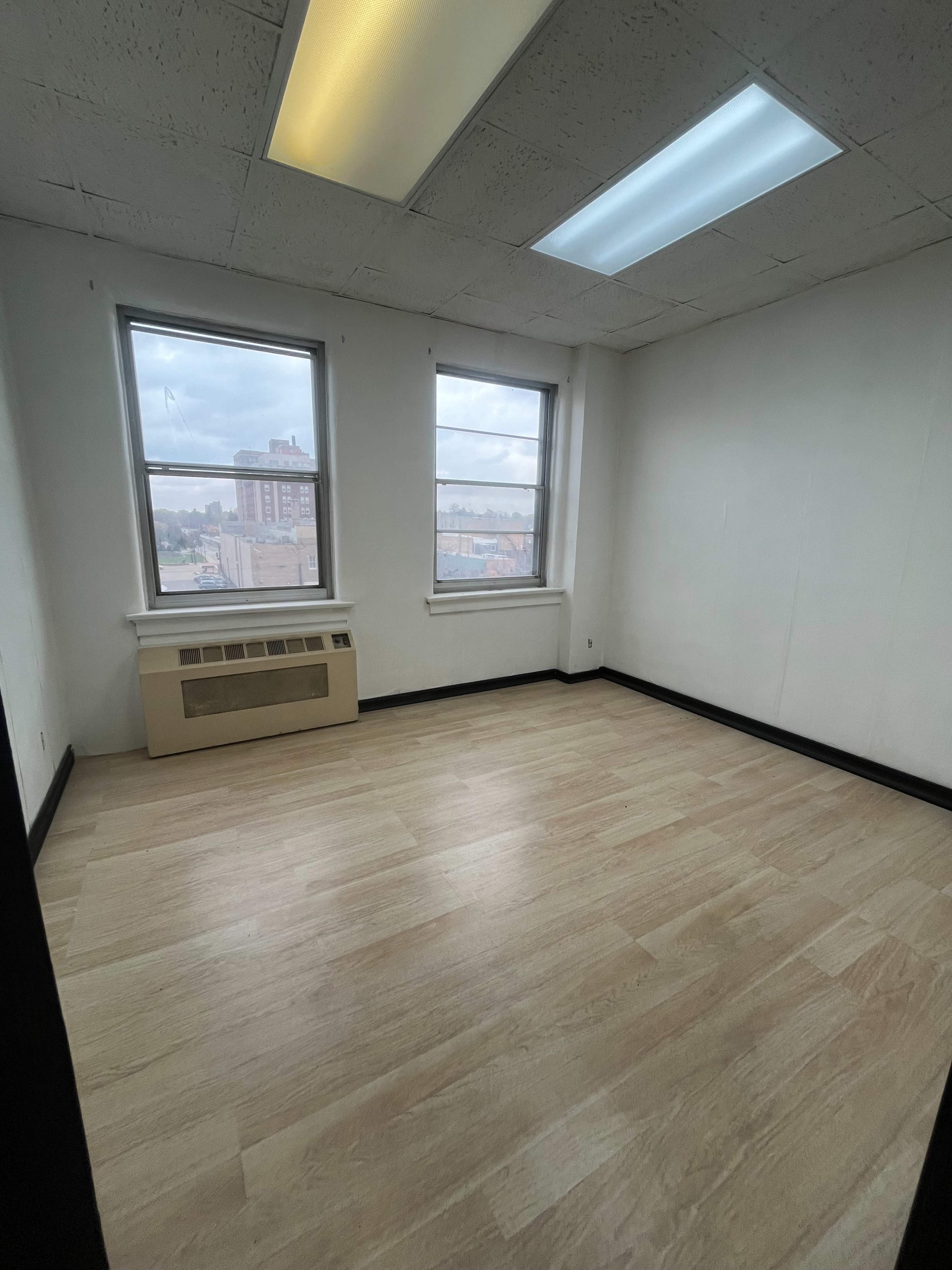 The image shows an empty room with two windows, a heating unit along one wall, and light-colored flooring.