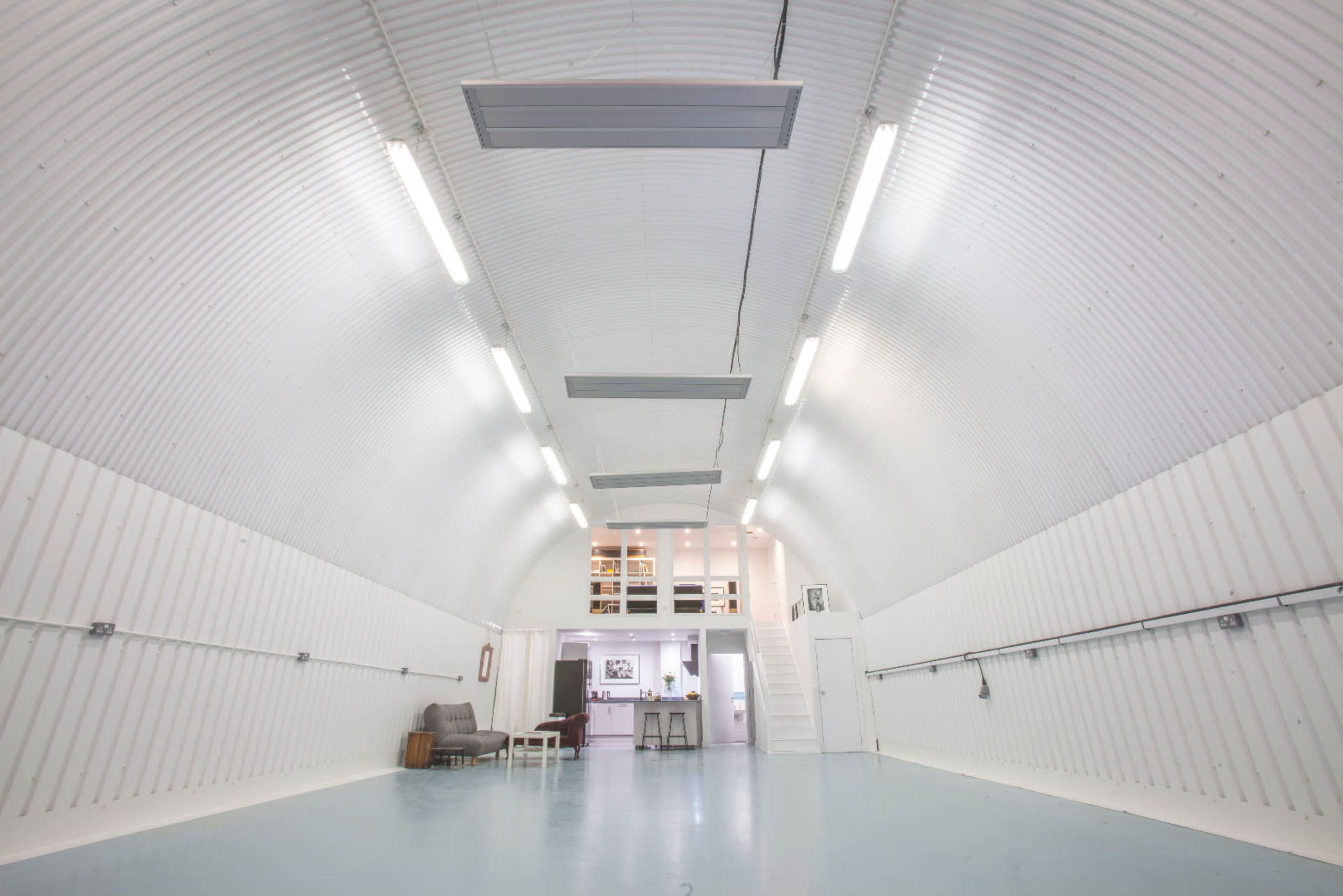 The image shows a spacious, white, corrugated interior with a high arch, featuring industrial-style lighting and a small multi-level area at the far end.