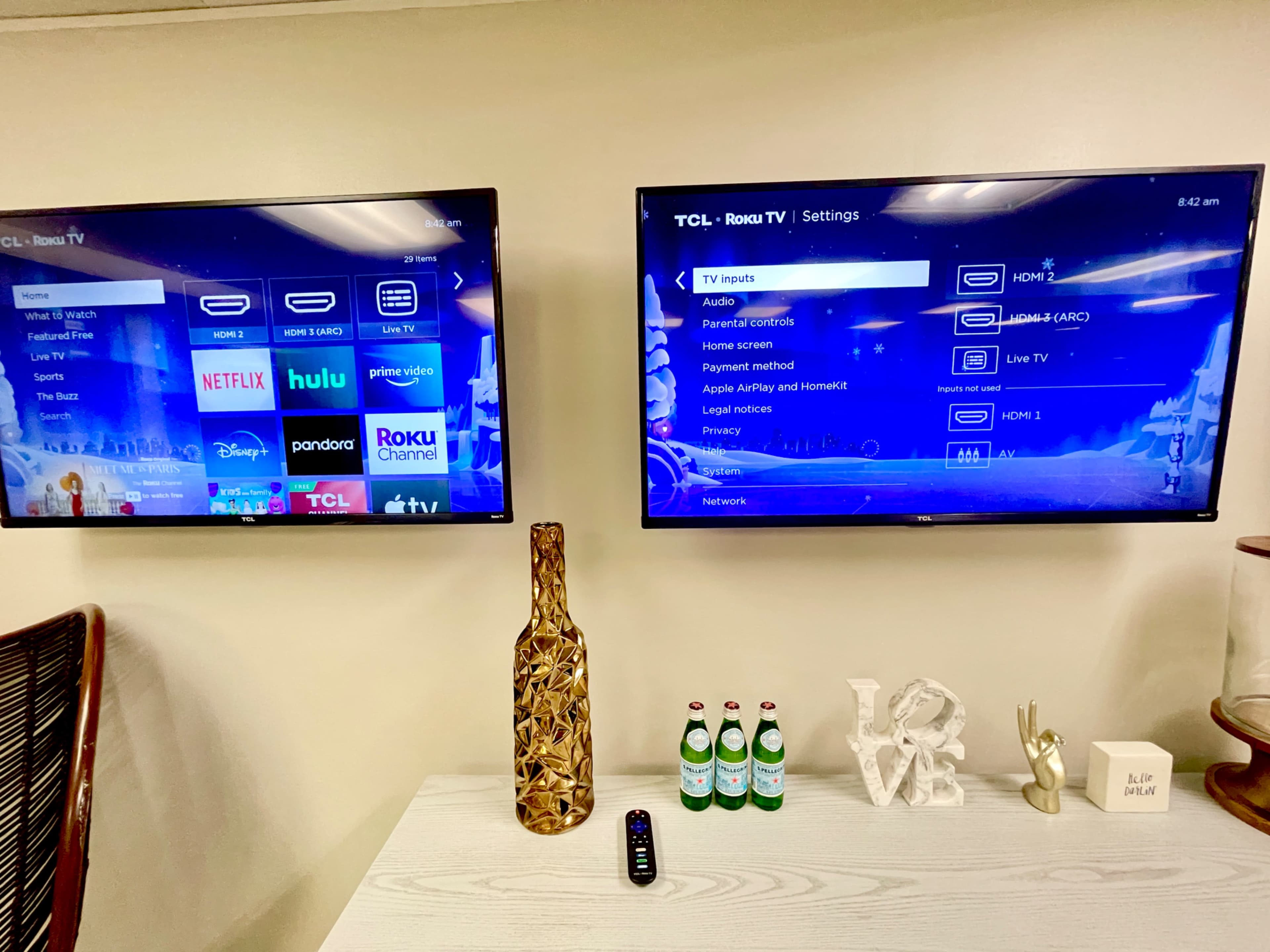 Two TCL Roku TVs mounted on a wall displaying various streaming service options, alongside a decorative vase and a few water bottles on a table.