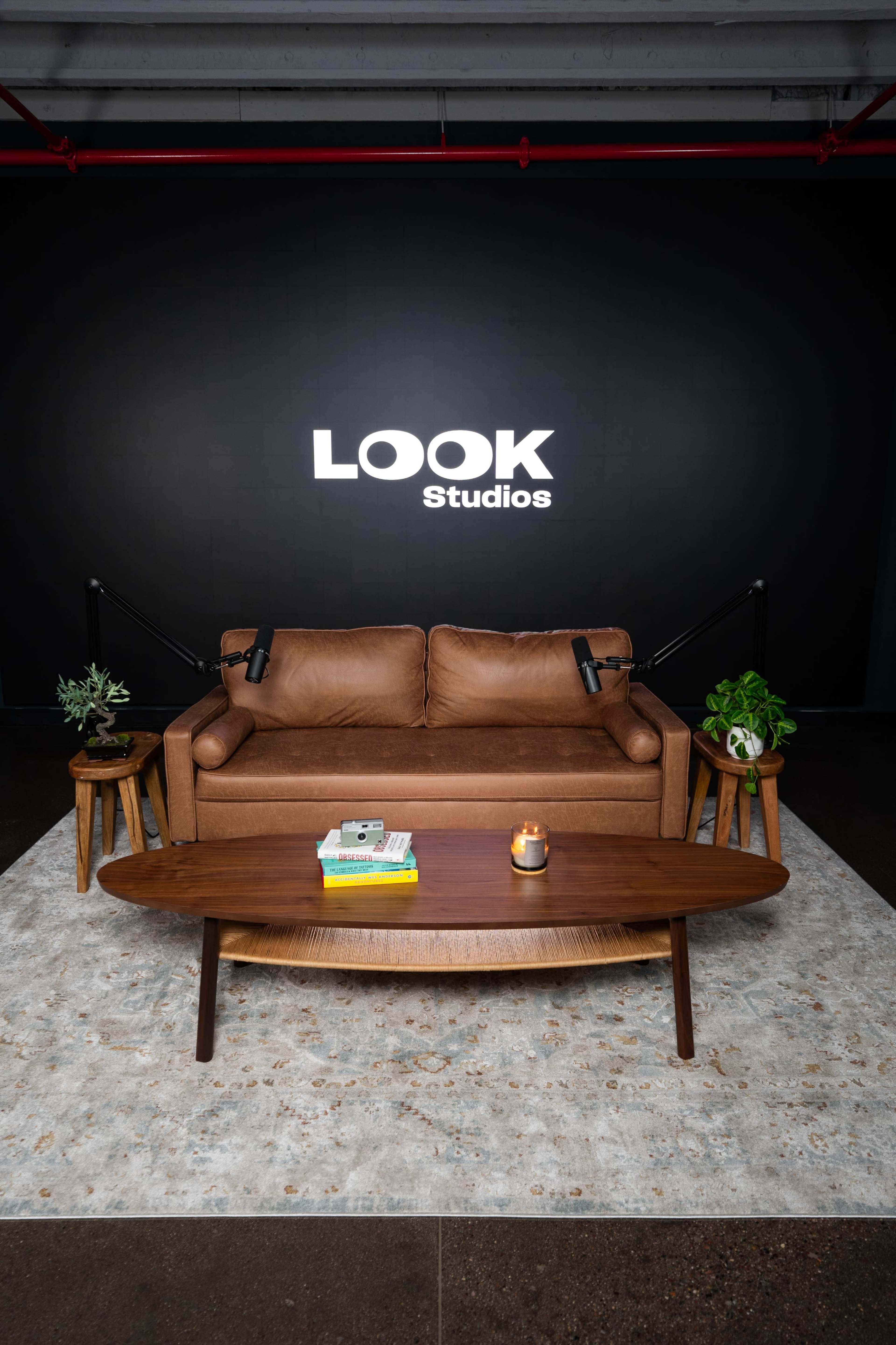A brown leather sofa is positioned in front of a dark wall featuring the words "LOOK Studios," accompanied by a wooden coffee table and plants on either side.