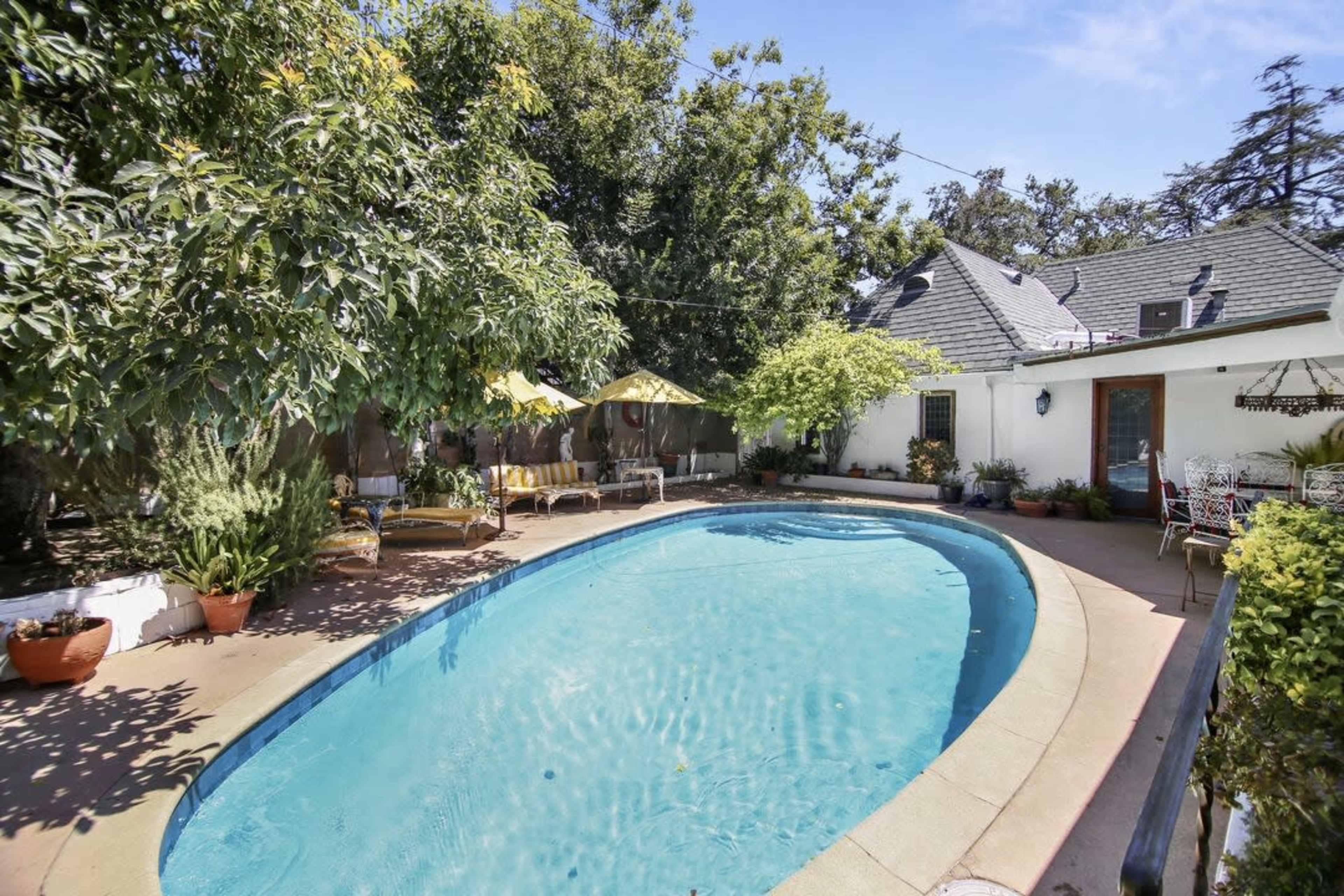 The image shows a swimming pool surrounded by a patio, lounge chairs, and greenery near a house.