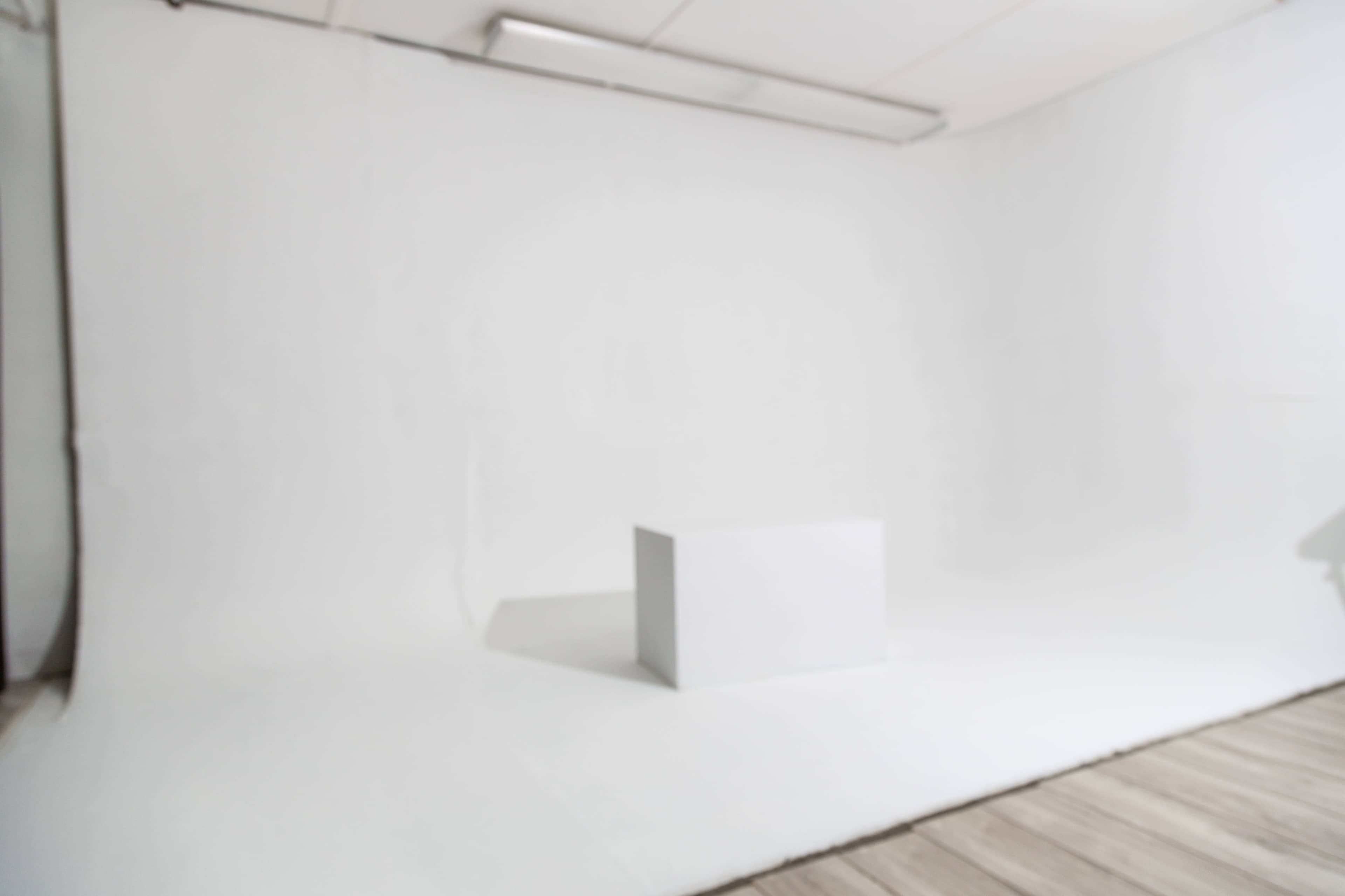The image shows a minimalistic studio space with a large white backdrop and a plain white pedestal in the center.