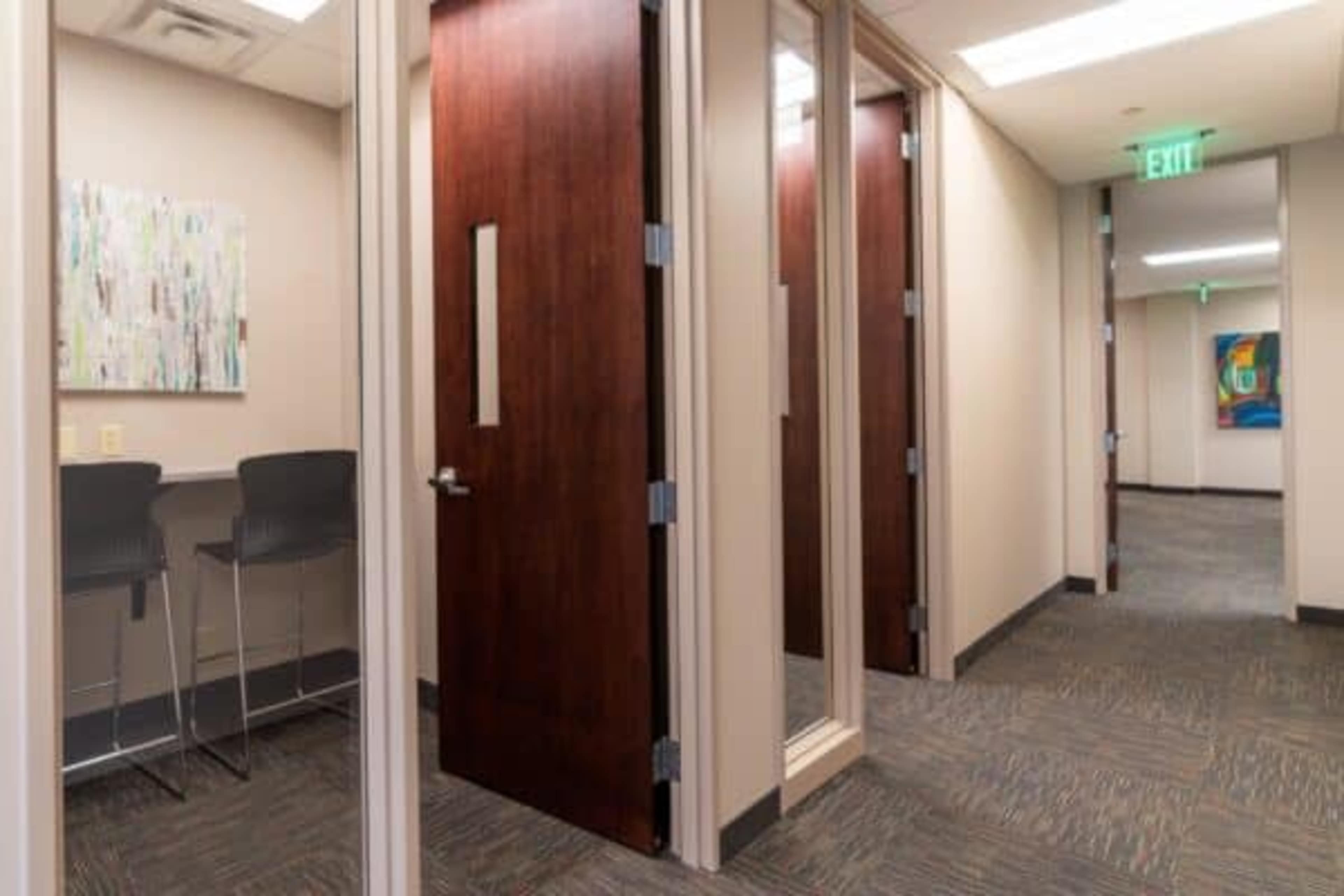 The image shows a hallway with multiple office doors, a small seating area with high chairs, and a colorful abstract painting on the wall.