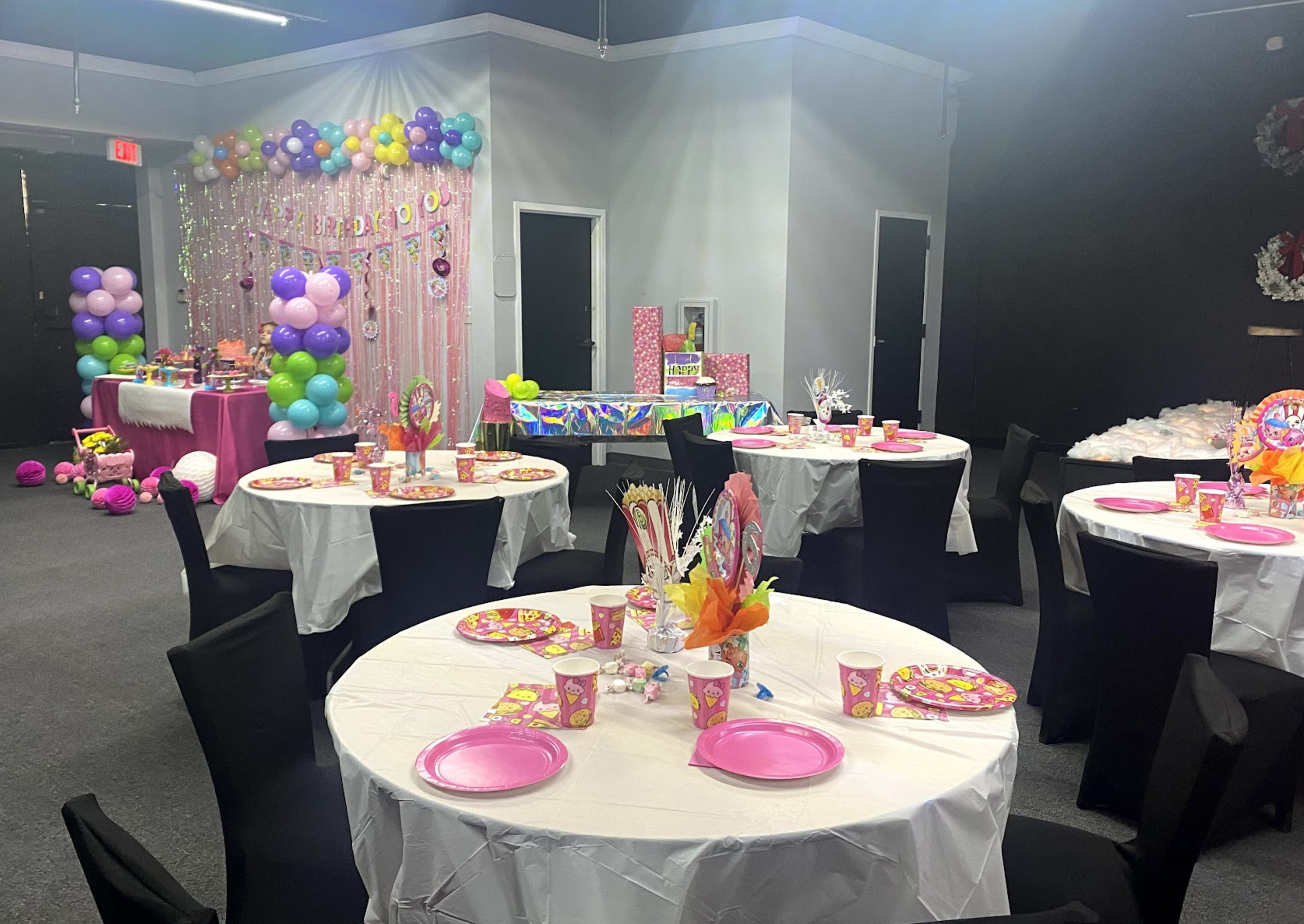 A decorated party venue with round tables covered in white linens, each set with pink plates and colorful party supplies, and vibrant decorations including balloons and a backdrop.