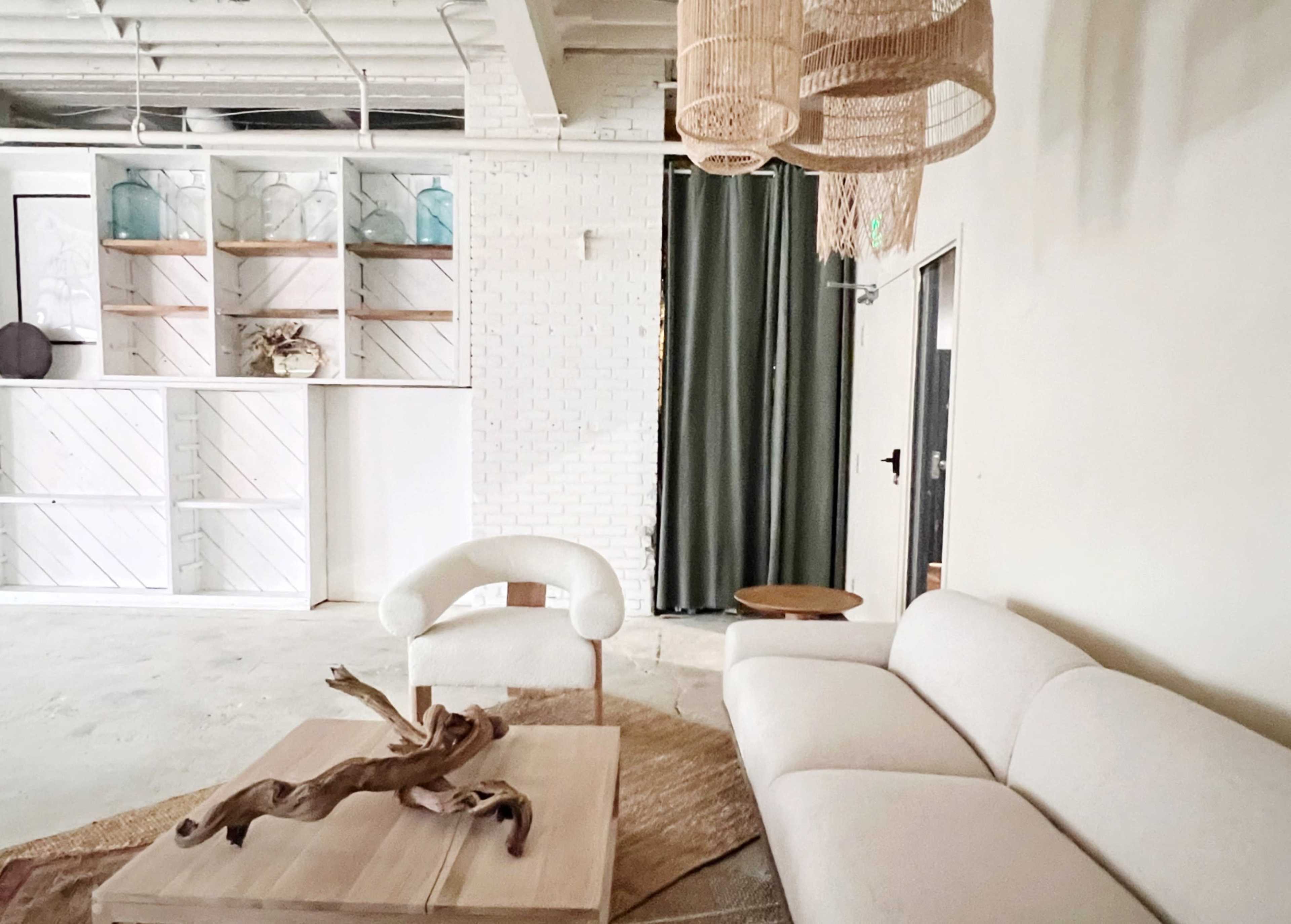 The image shows a minimalist interior living space featuring a white sofa, a wooden coffee table with a decorative element, and a green curtain in front of a light-colored wall.