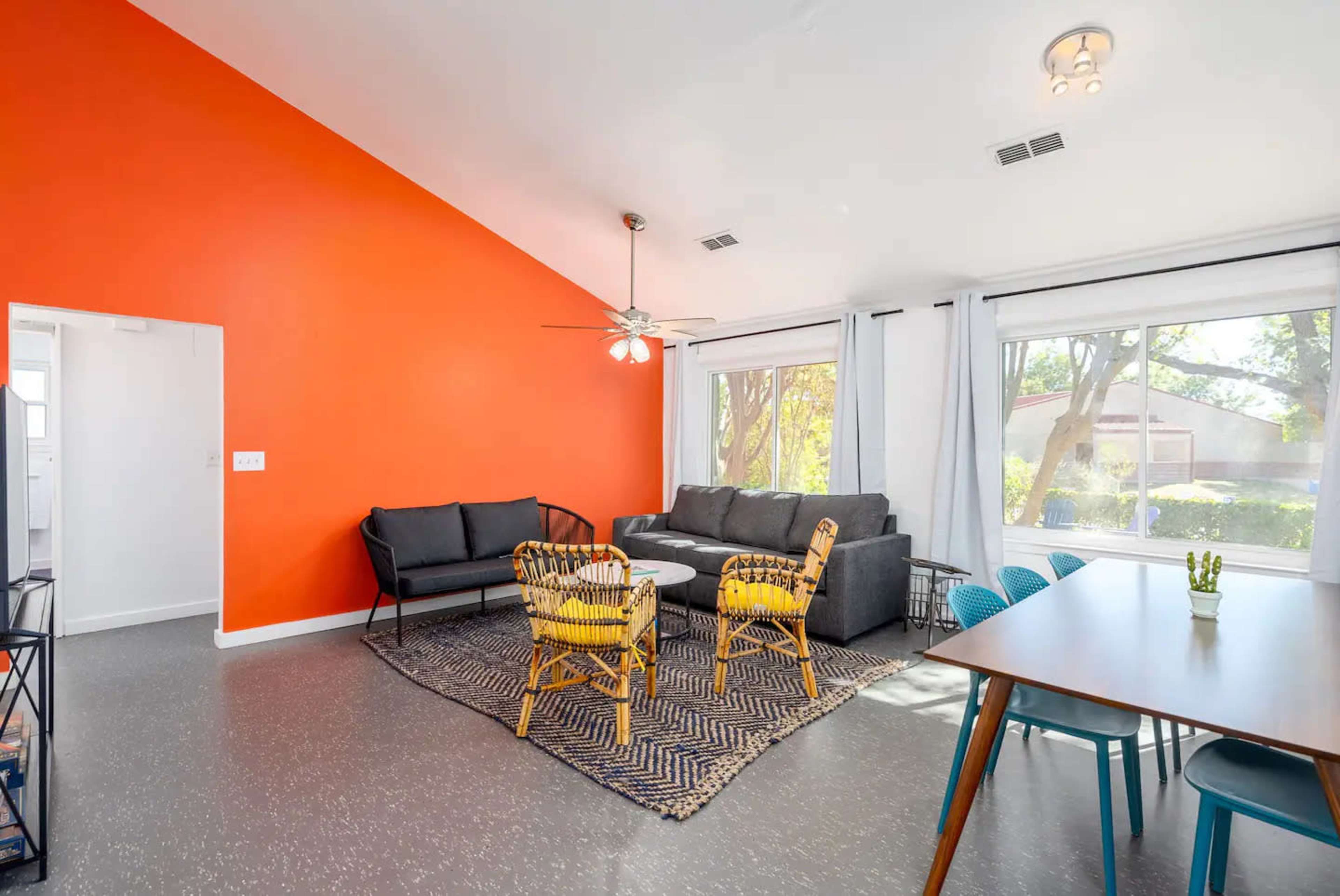 The image shows a bright living space with an orange accent wall, featuring a gray sofa, a patterned rug, wicker chairs, and a dining table with teal chairs.