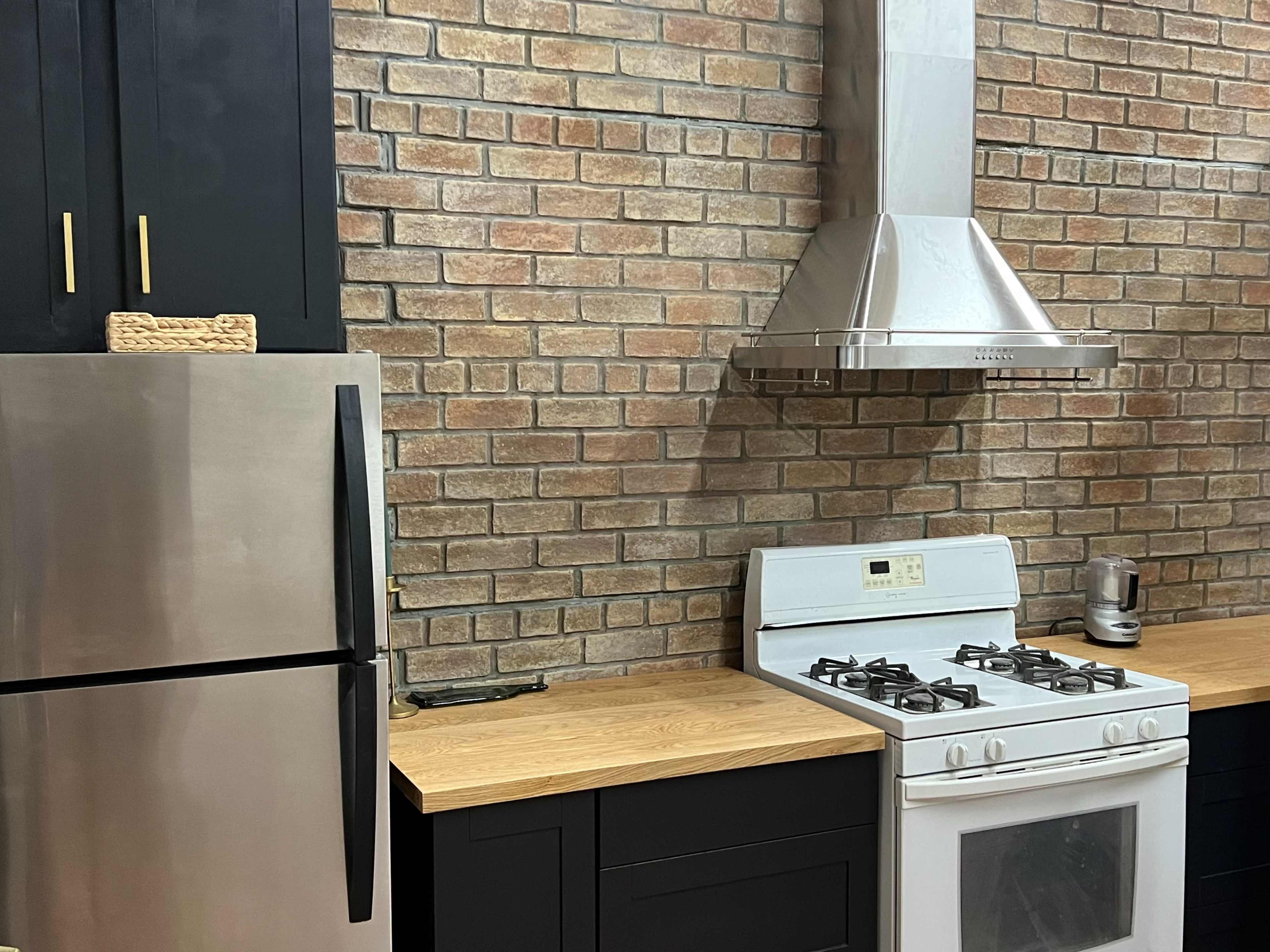 A kitchen with a stainless steel refrigerator, a gas stove, and a hood vent above, set against a brick wall and wooden countertops.