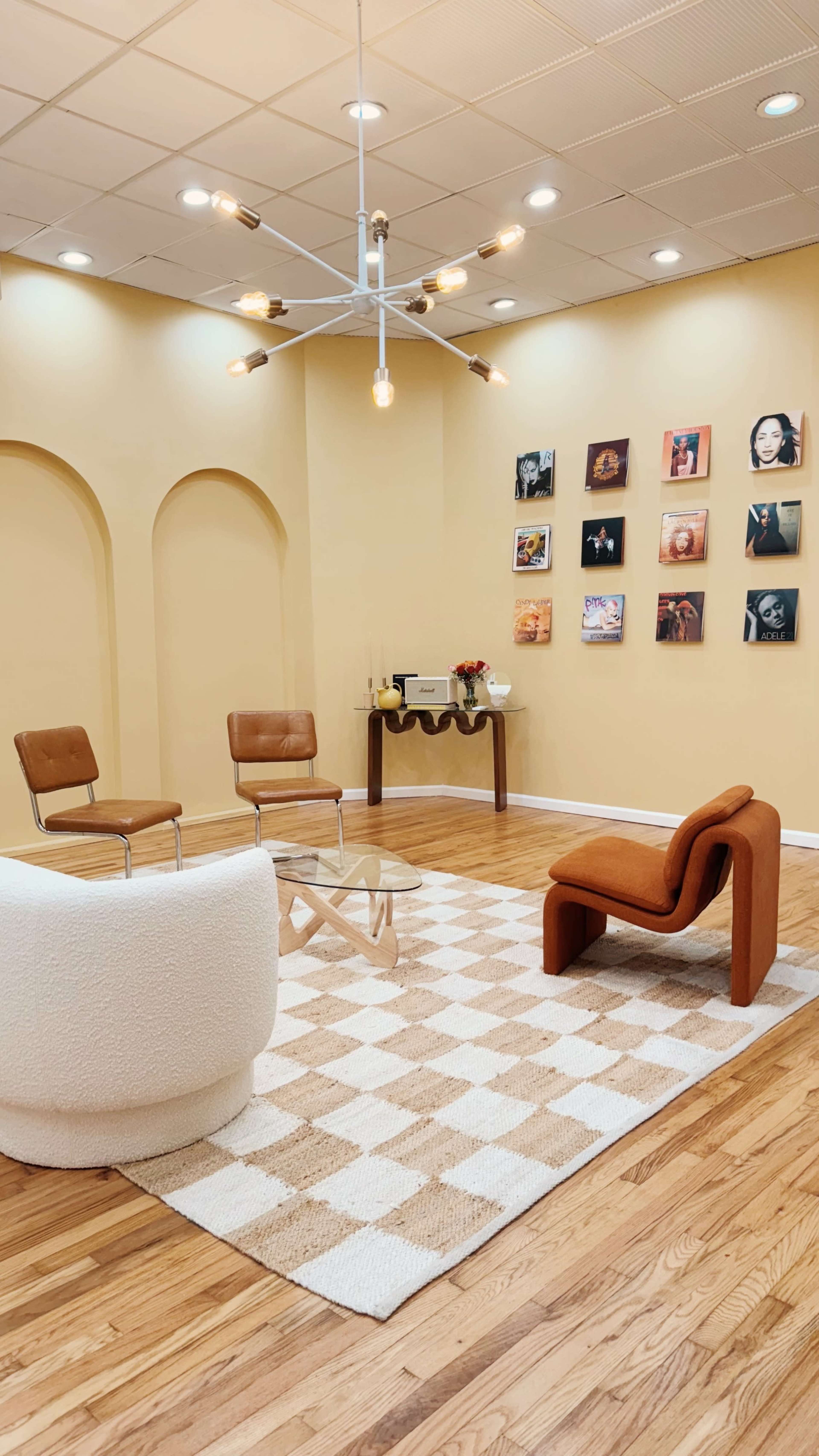 The scene features a minimalist lounge area with two brown chairs, an orange couch, a patterned rug, and a collection of framed photos on the wall.