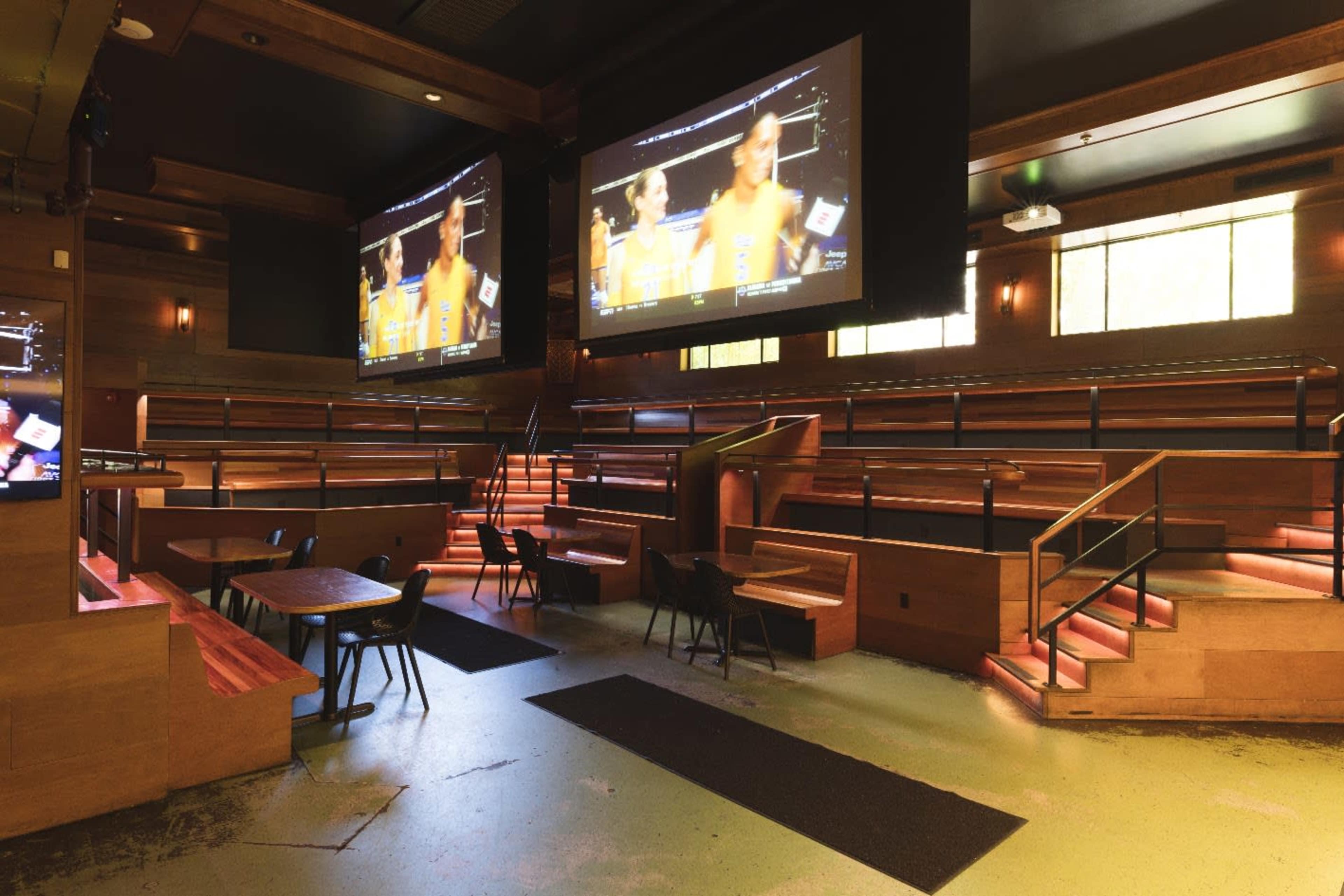 The image shows a spacious indoor venue with wooden seating, two large screens displaying a sports event, and illuminated stairs.
