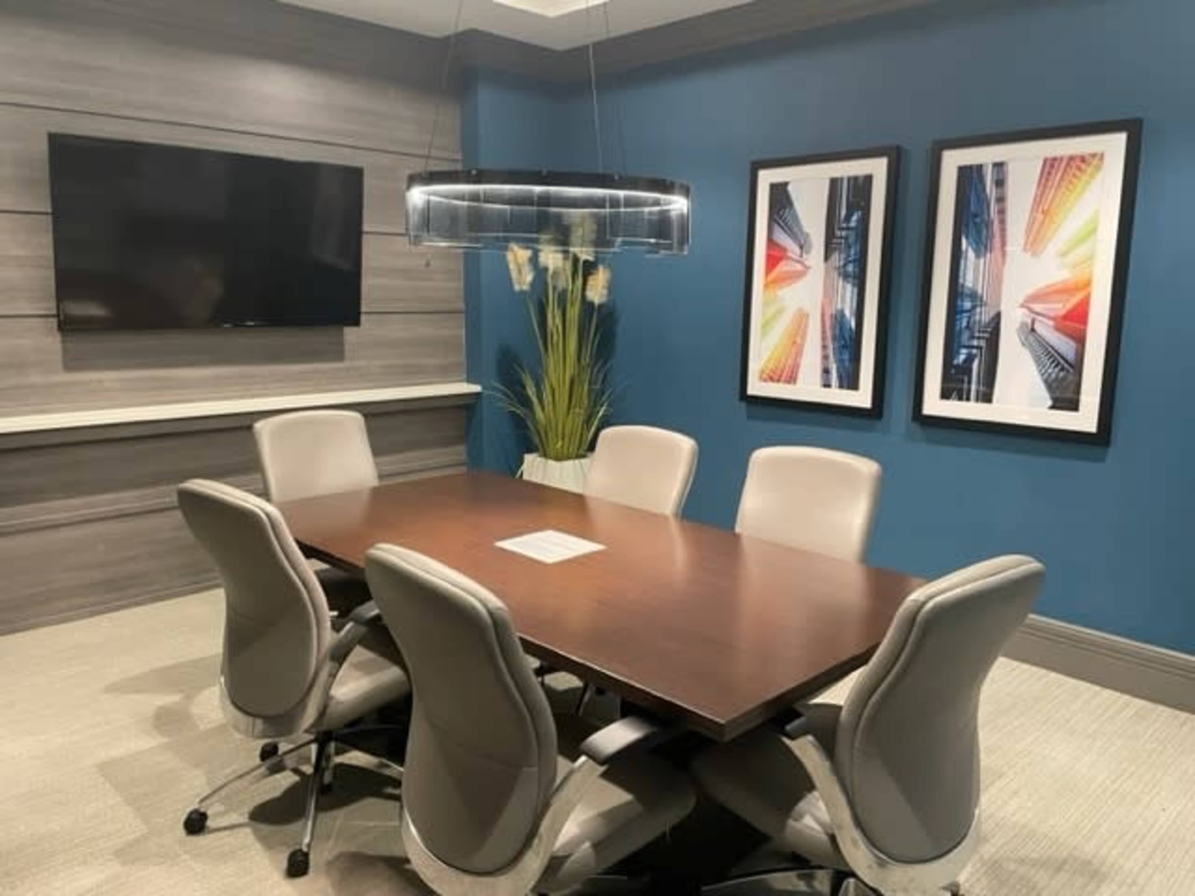 A modern conference room features a long wooden table surrounded by six ergonomic chairs, with a television and two framed art pieces on the walls.
