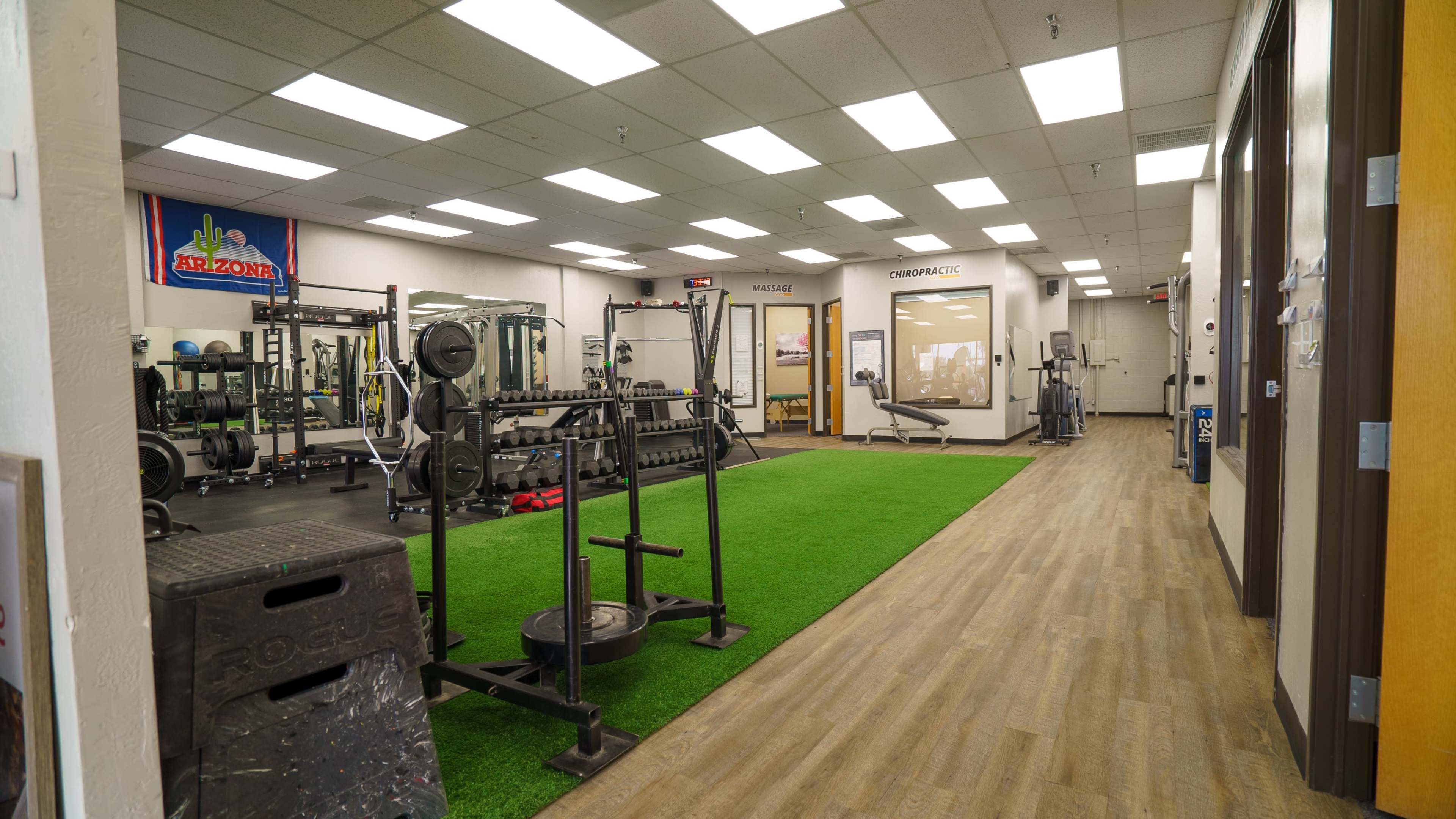 The image shows a well-equipped gym with weightlifting stations, artificial turf, and fitness machines lining the walls.