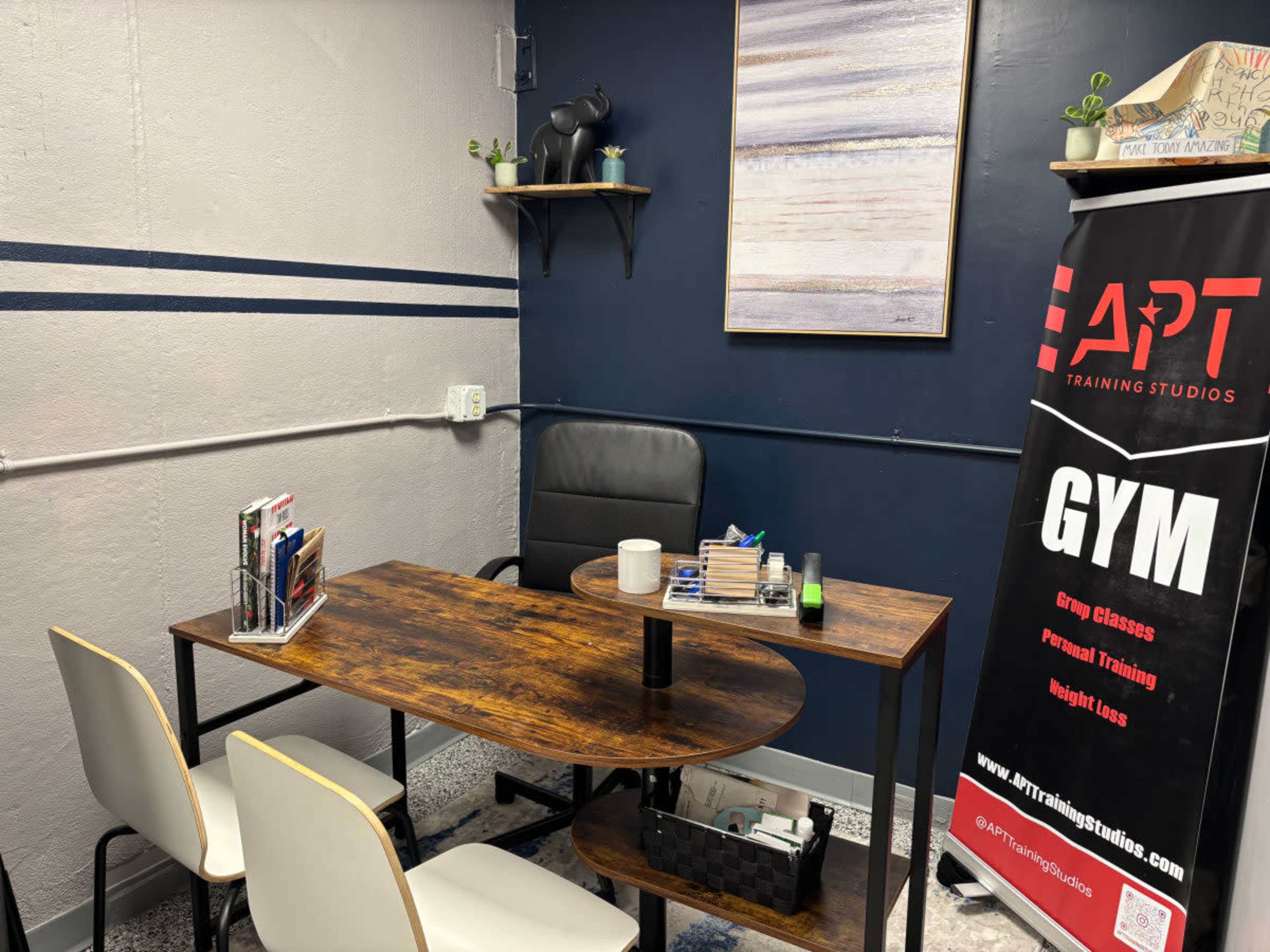 The image shows a small office or training studio with a wooden desk, a black chair, a bookshelf, and a banner advertising gym services.