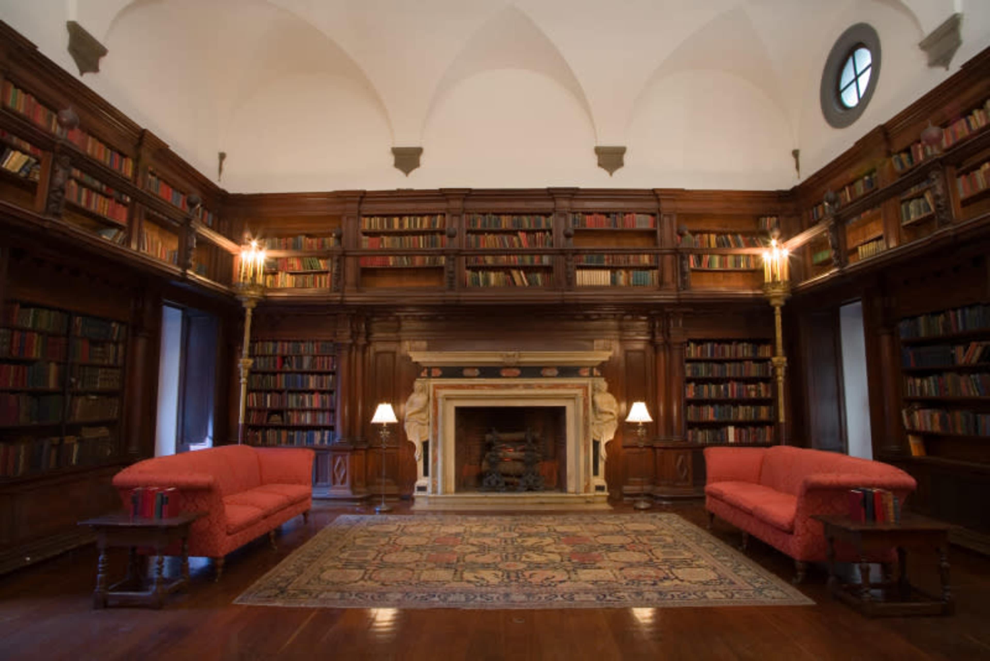 The image shows a spacious library with wooden shelves filled with books, two red sofas, and a fireplace framed by a decorative mantel.