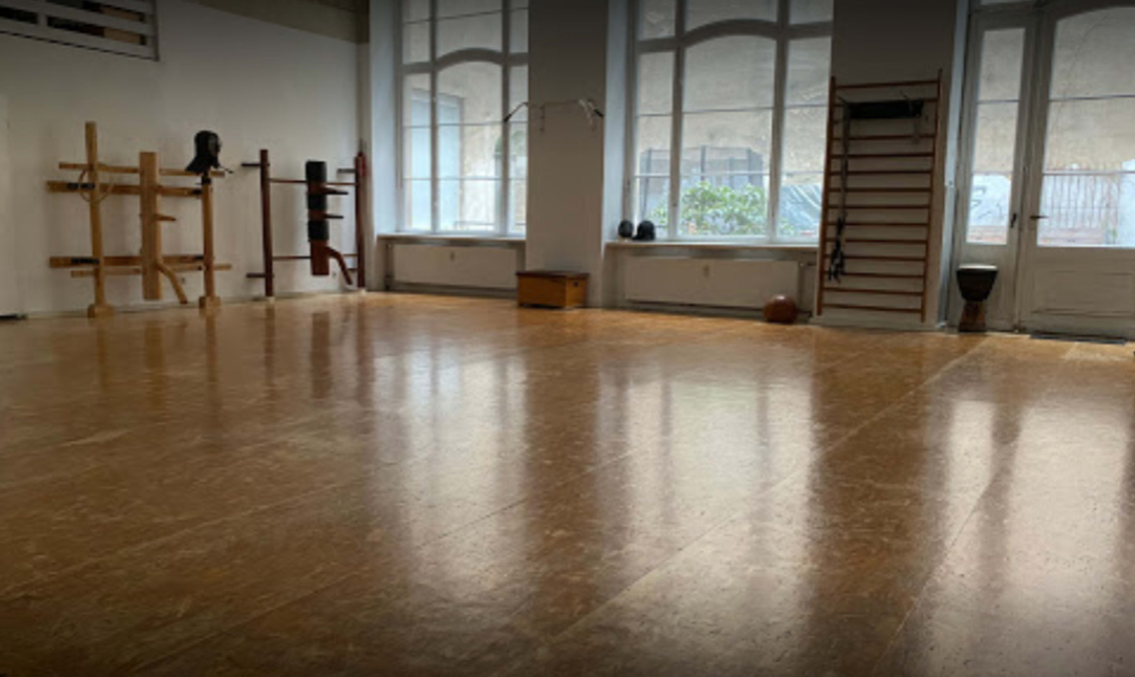 The image shows a spacious, empty room with wooden flooring and various martial arts training equipment along the walls.