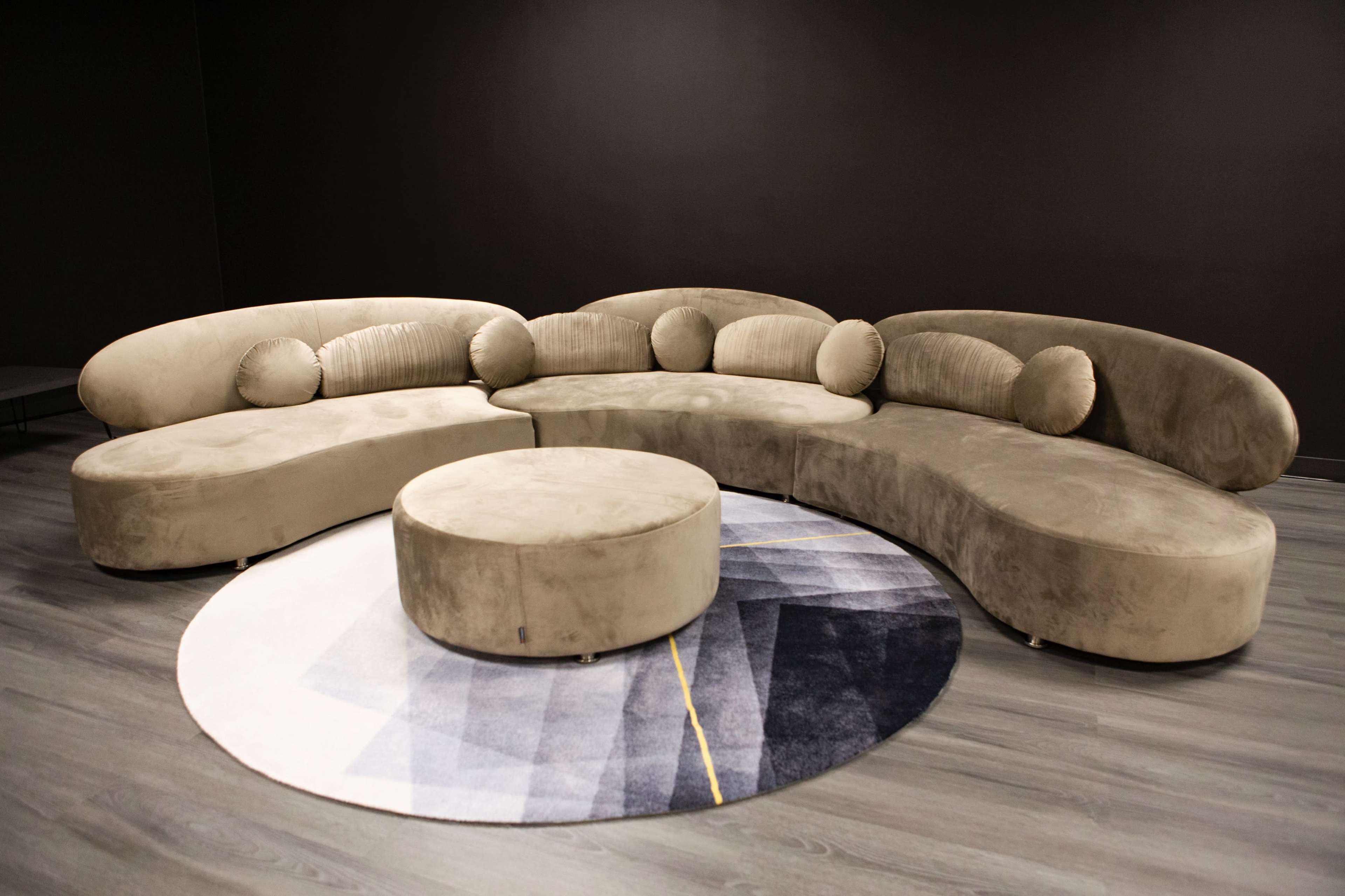 A curved beige sofa with circular cushions is arranged around a round, patterned geometric rug on a dark floor.