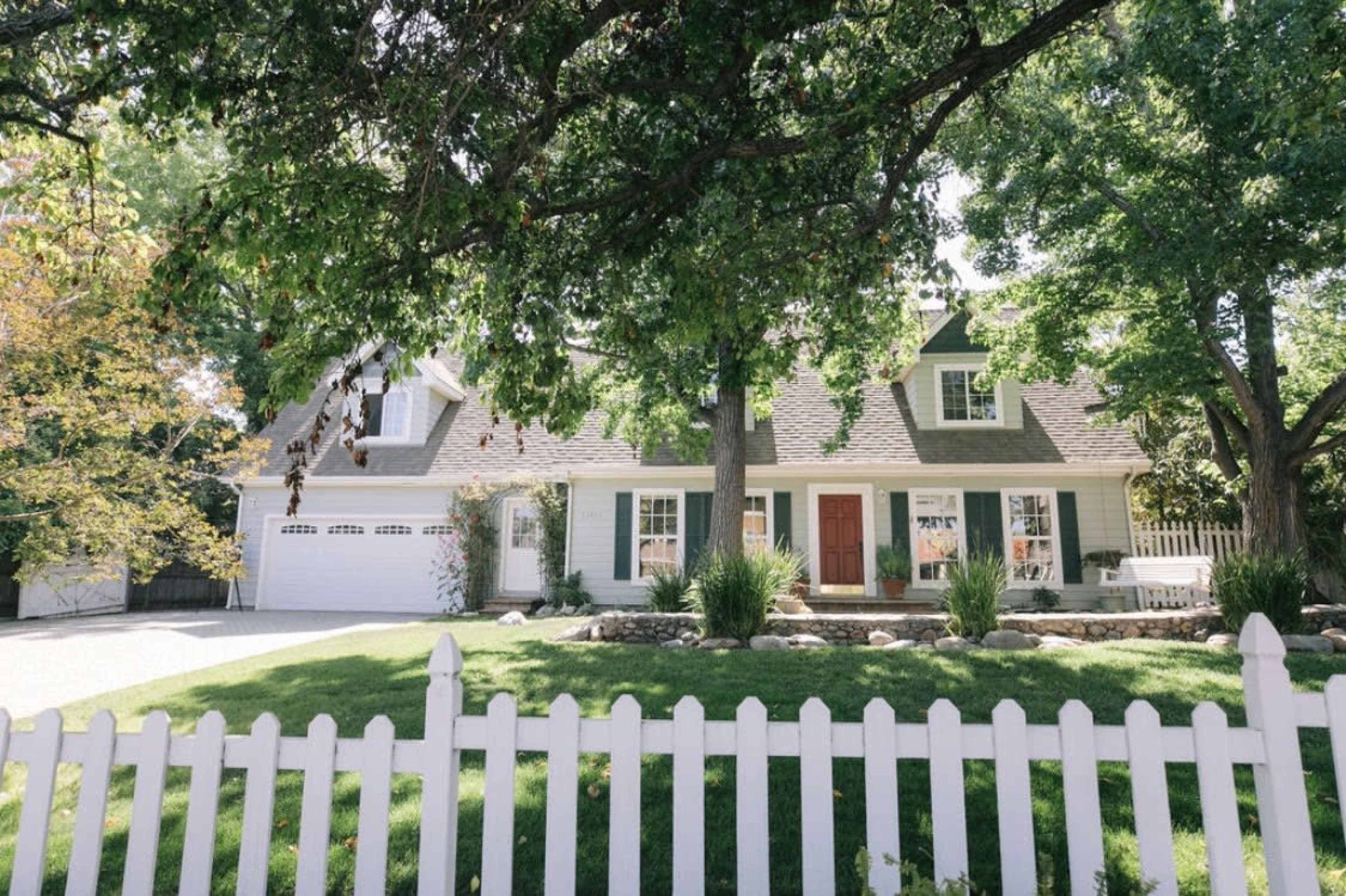 A white picket fence surrounds a green lawn in front of a two-story house with a sloped roof and a garage.