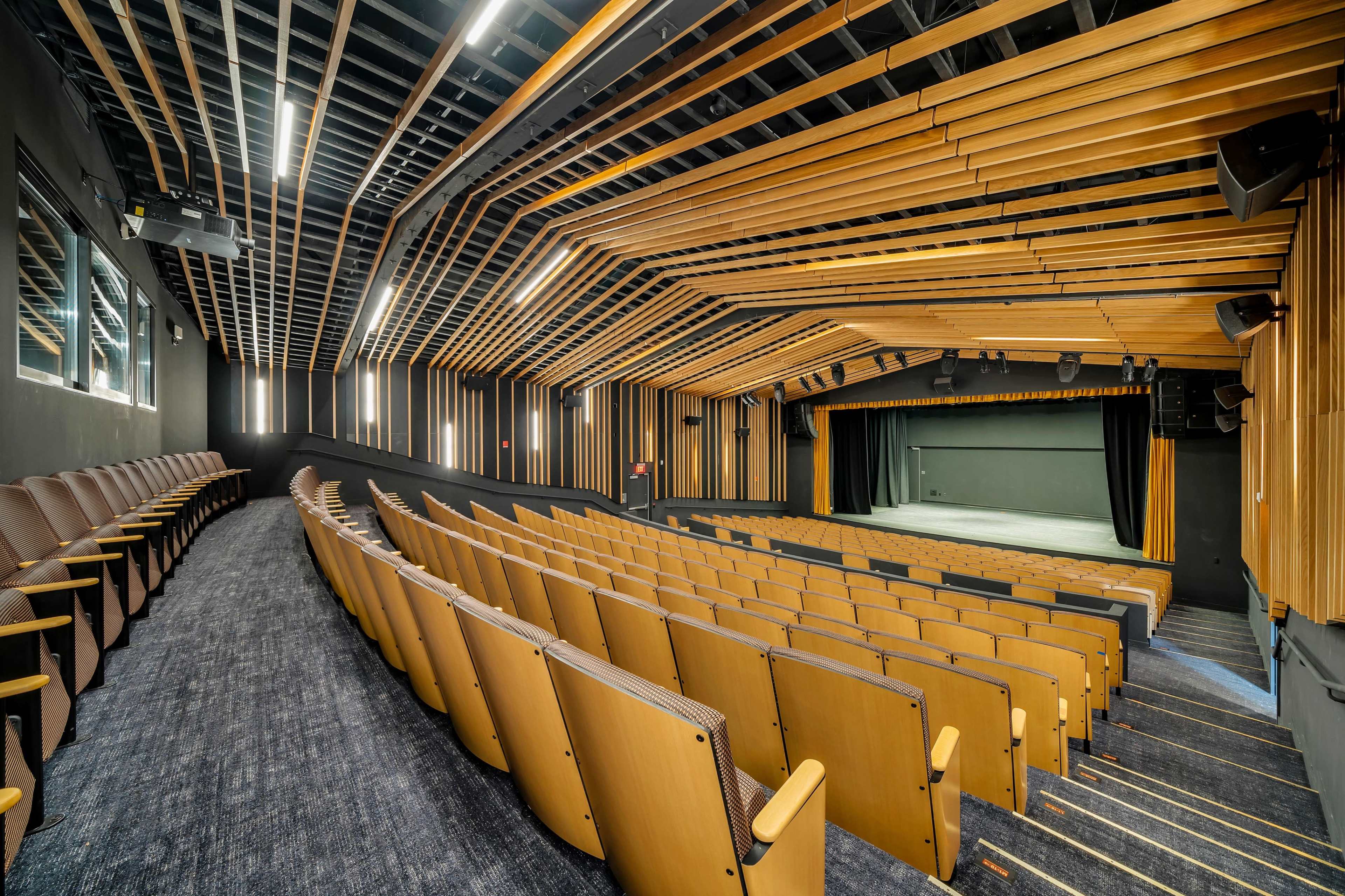 An interior view of a modern theater with wooden seating and a stage illuminated by overhead lighting.