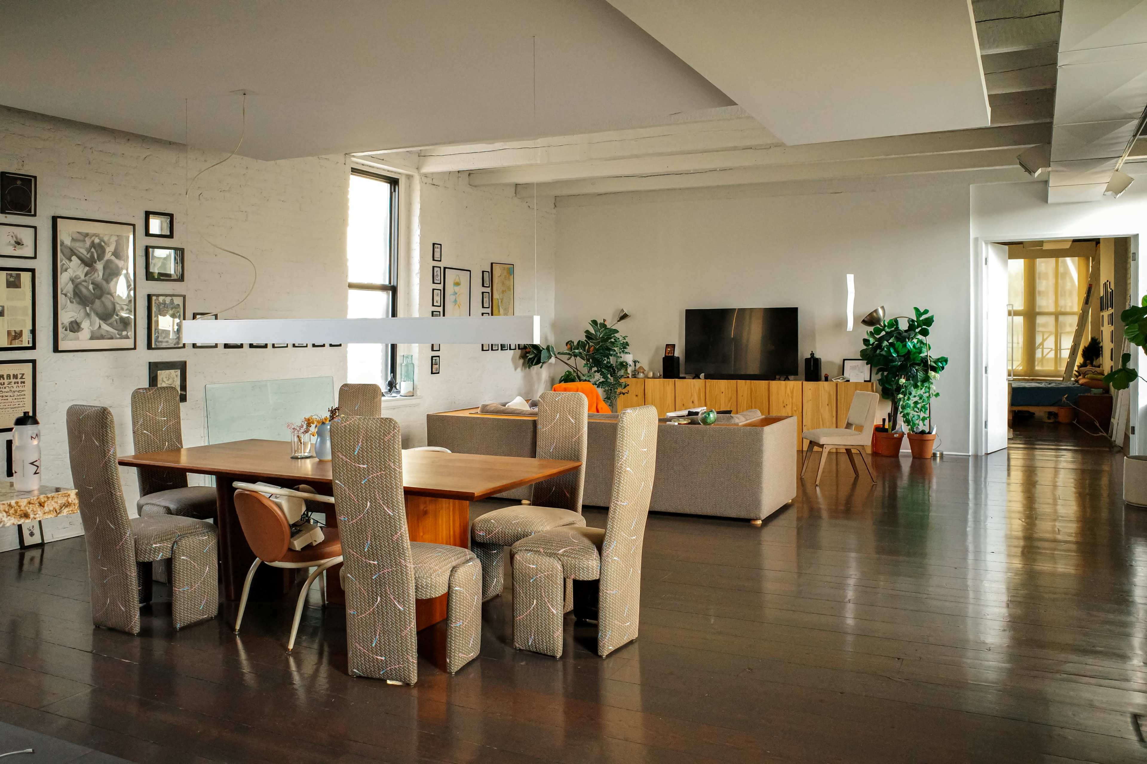 The image shows a spacious, modern living area featuring a dining table with upholstered chairs, a TV set on a wooden cabinet, and various plants around the room.
