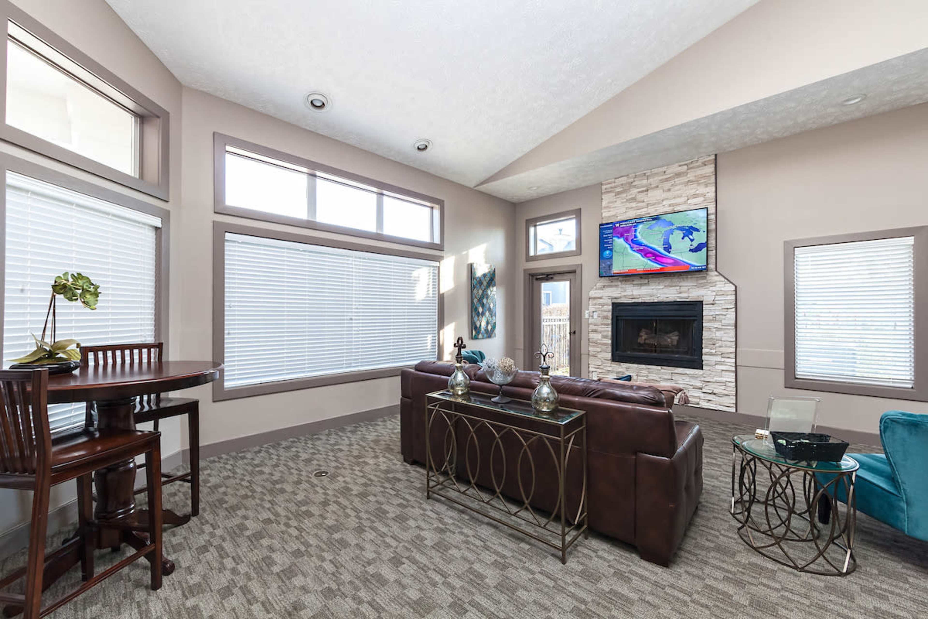 A modern living room with large windows, a stone fireplace, a television mounted on the wall, and a combination of seating and tables.