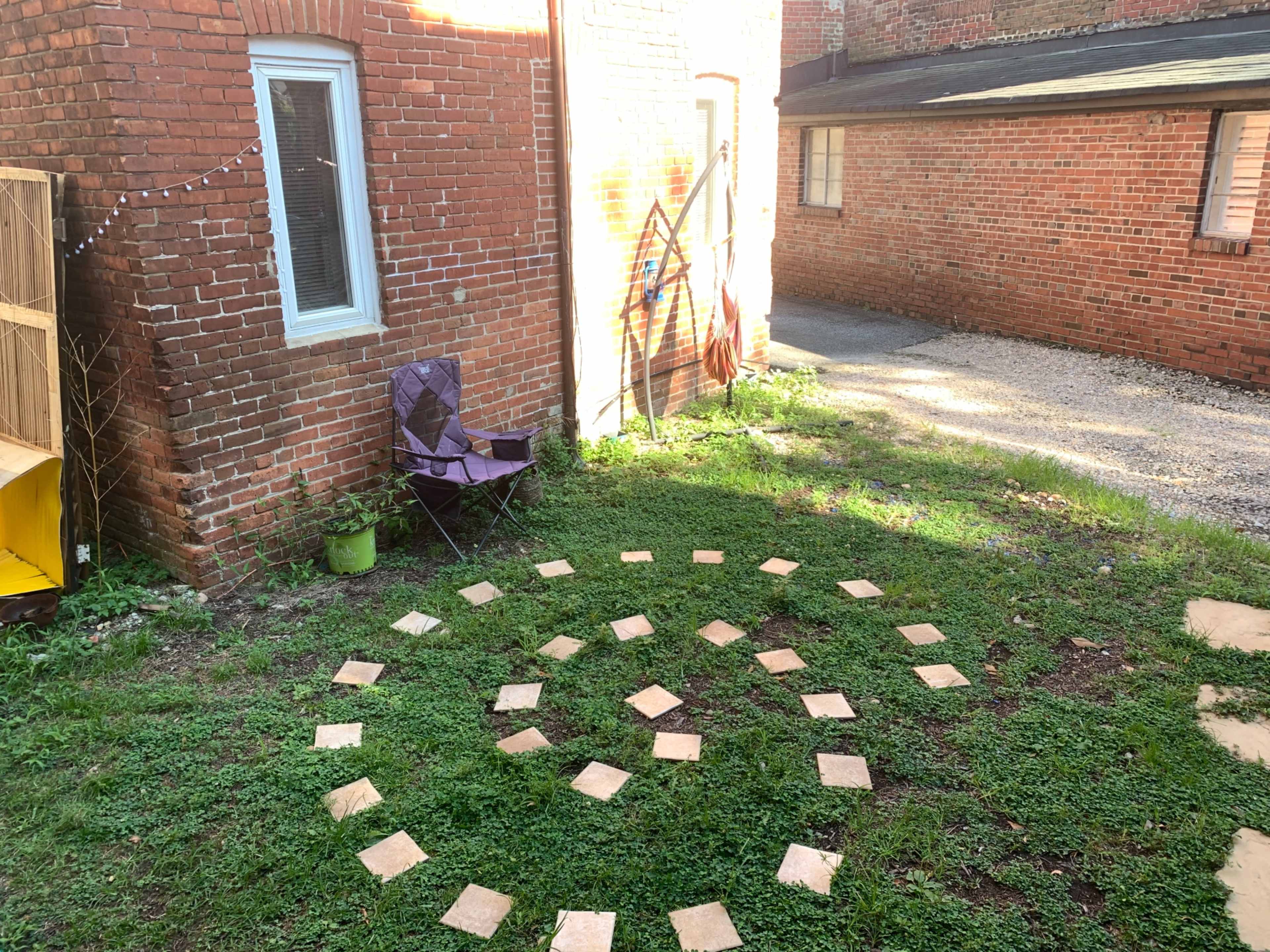 A grassy area with a spiral pattern made of square tiles surrounded by a purple folding chair and red brick buildings in the background.