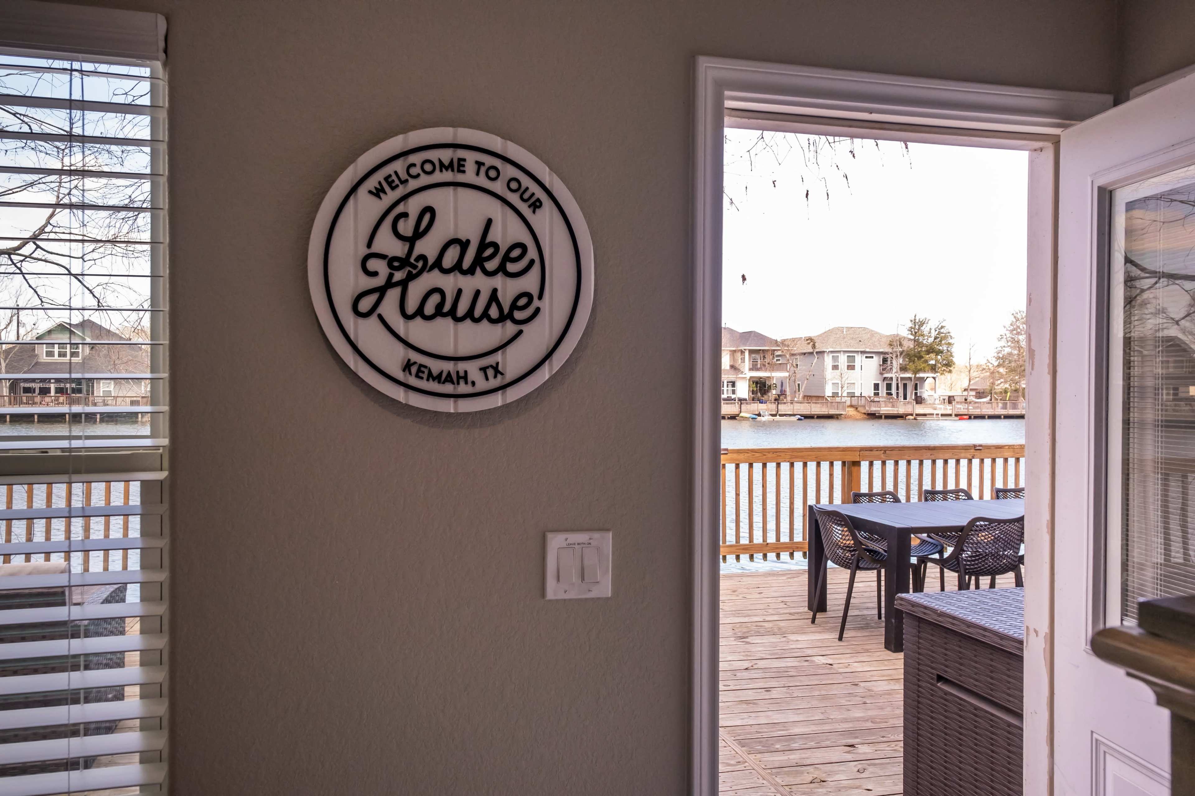 A welcome sign for a lake house is mounted on the wall, facing a view of a deck and the lake beyond.