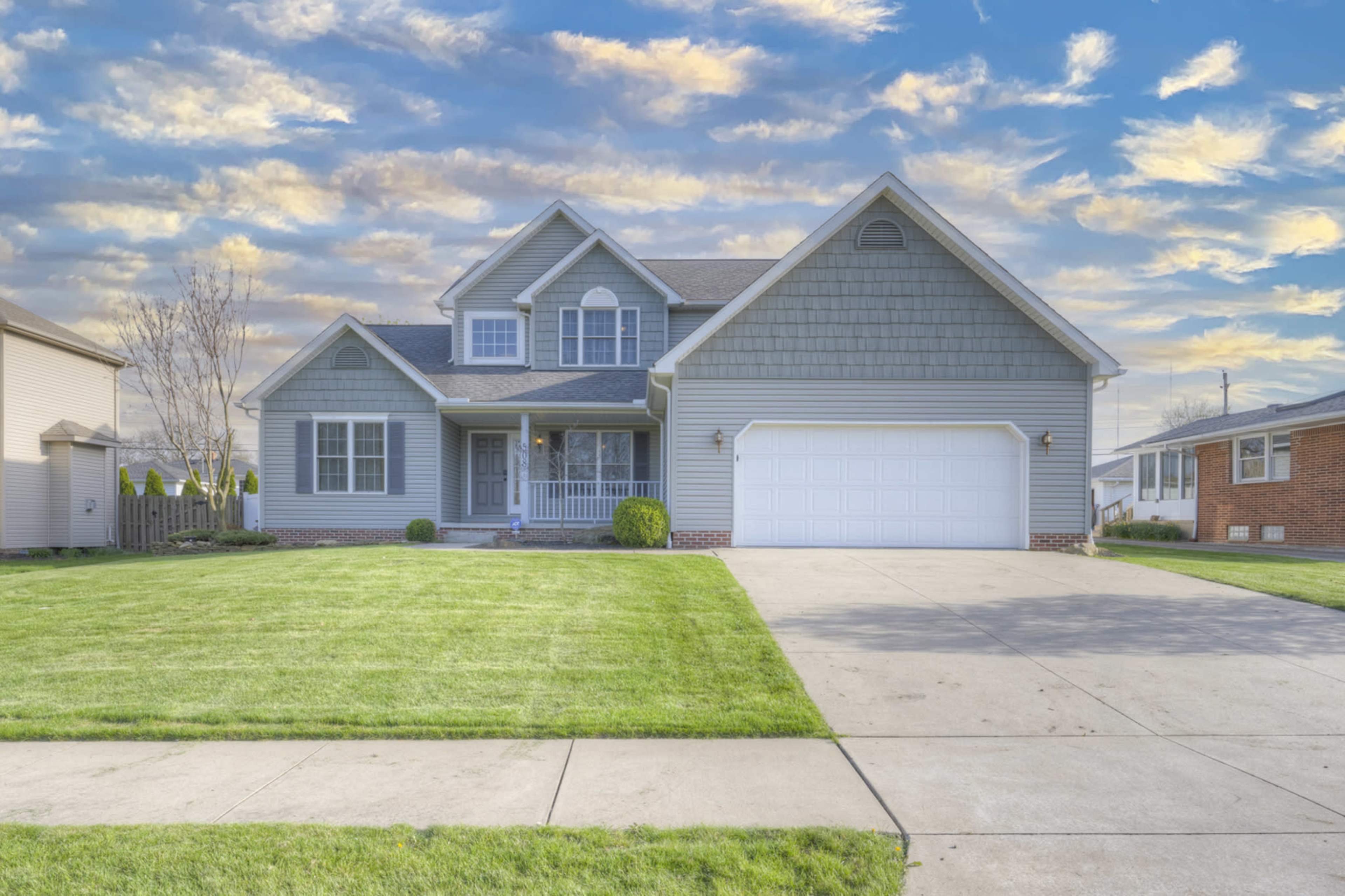 A gray two-story house with a front porch and a large driveway is set against a blue sky filled with clouds.