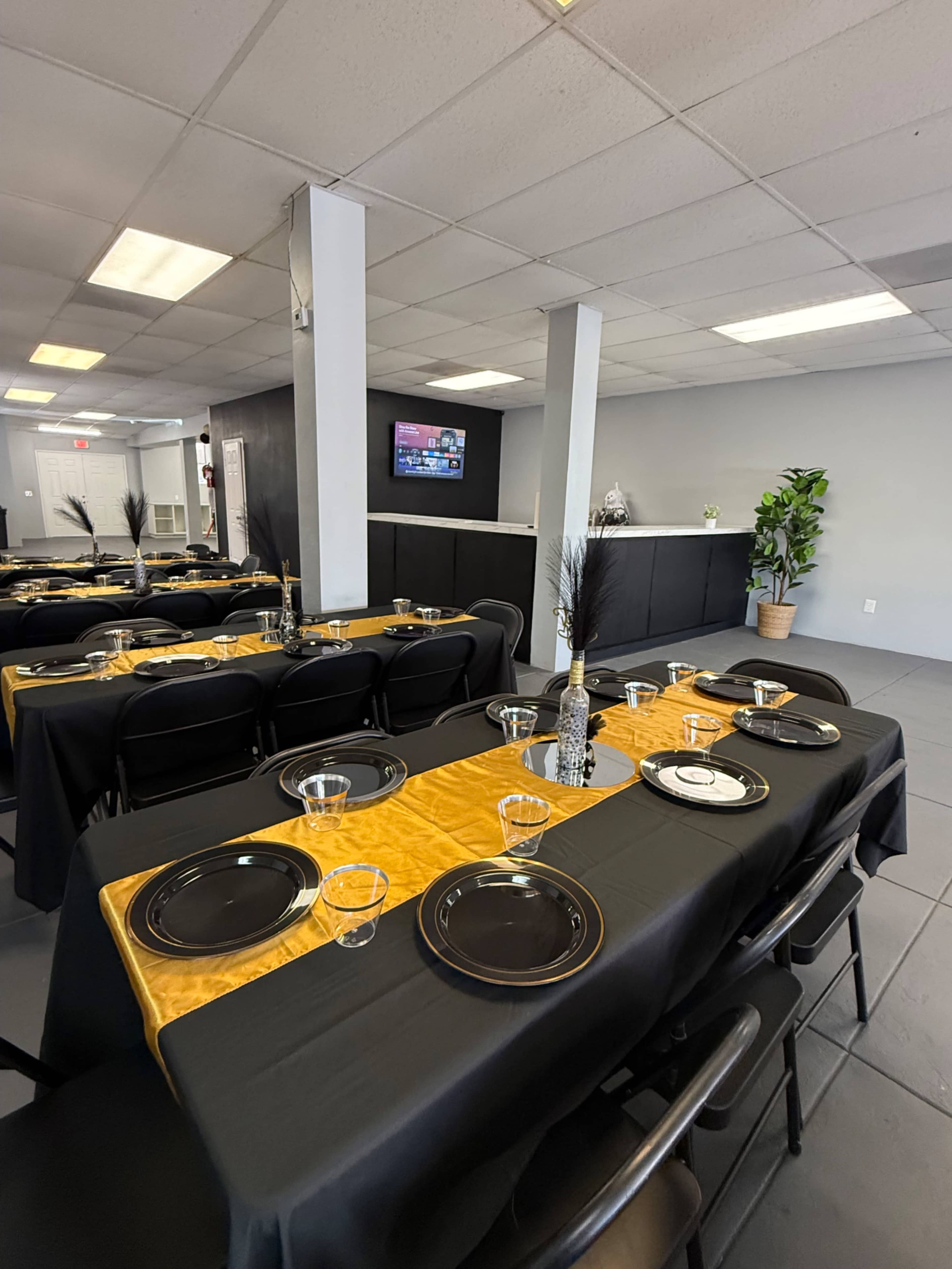 The image displays a dining area set up with several tables covered in black and gold tablecloths, arranged with plates and glassware, and featuring a large TV on the wall in the background.