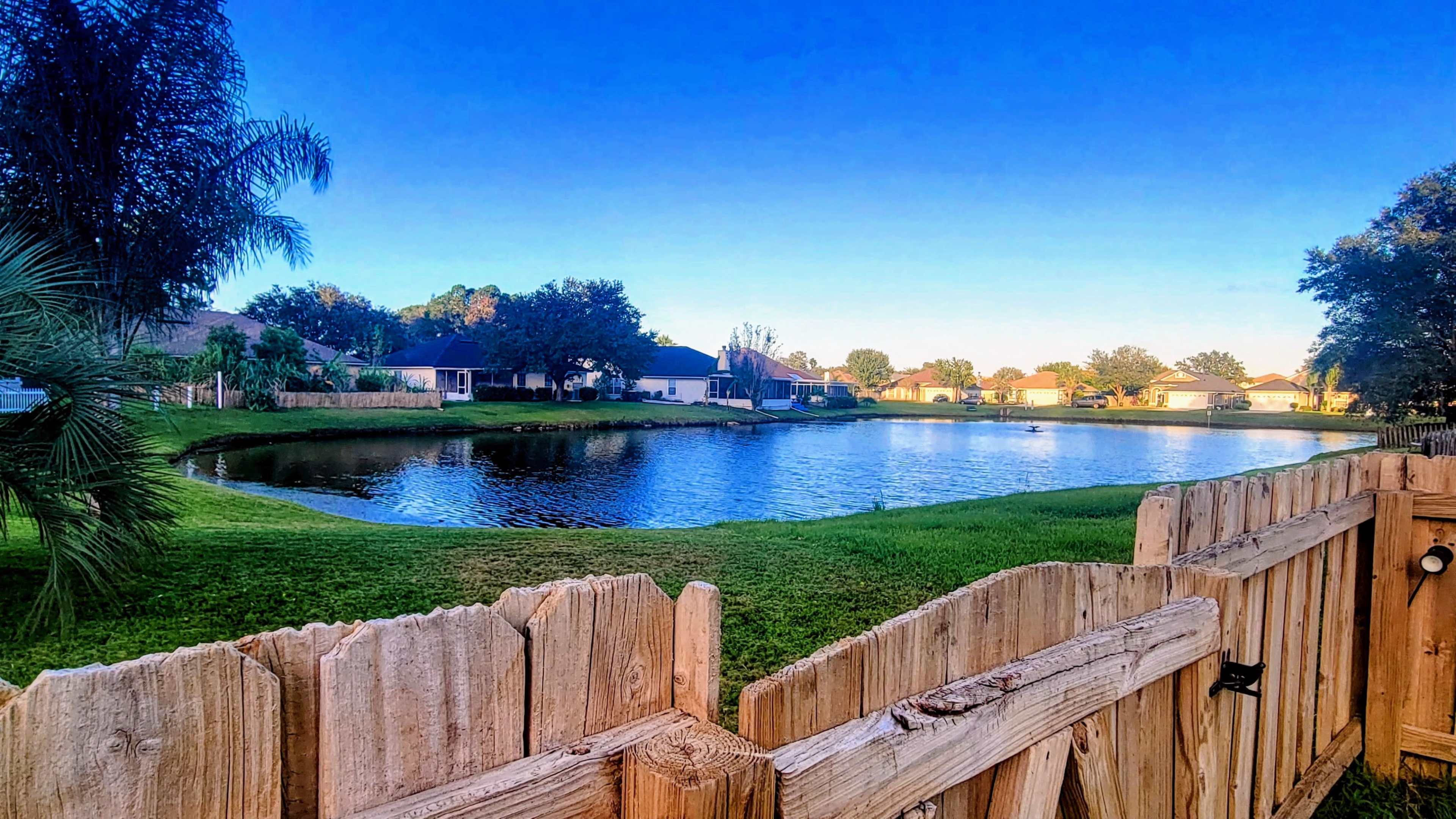 A tranquil pond surrounded by grassy areas and houses, with a wooden fence in the foreground.