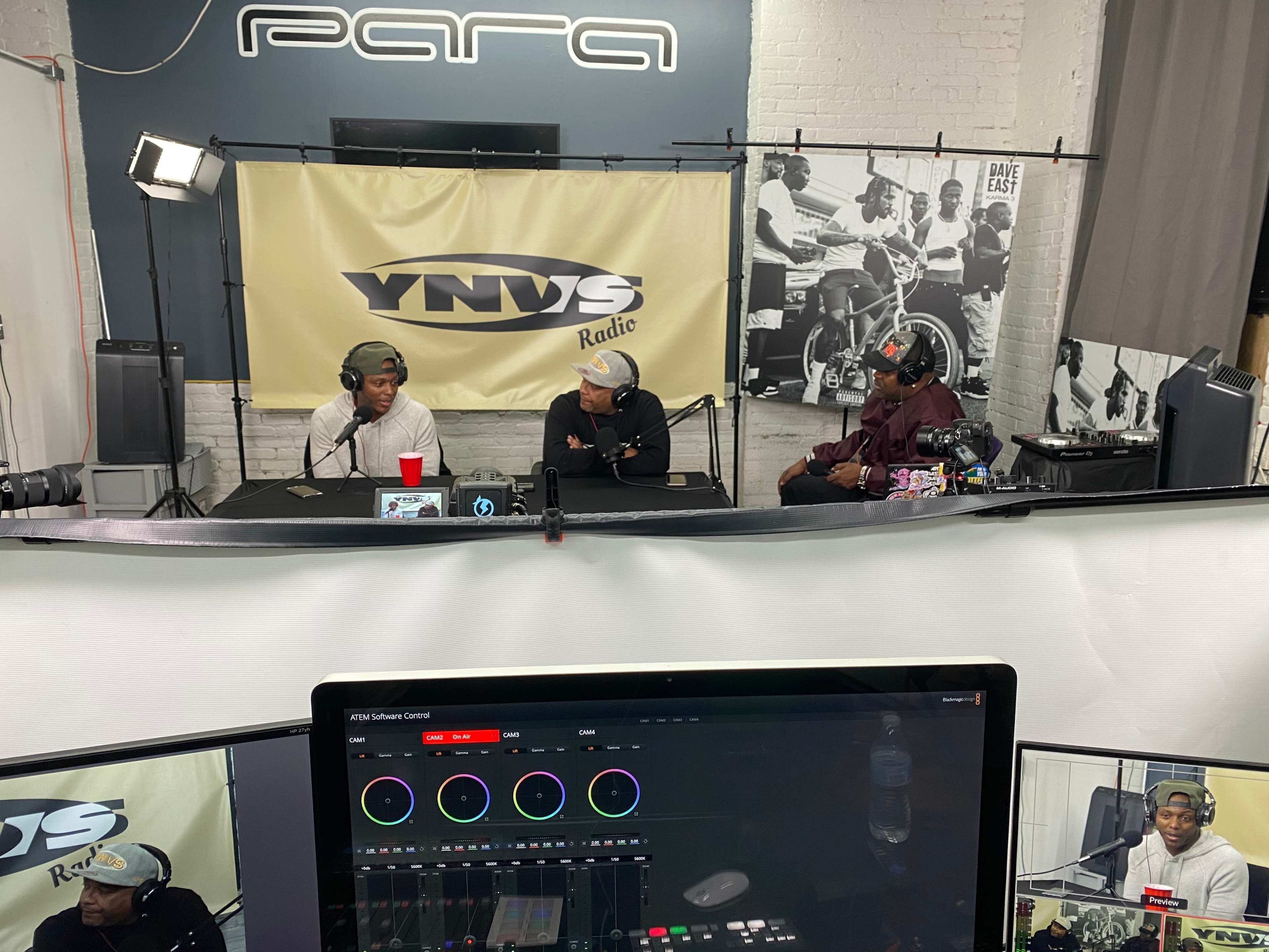 Three individuals sit at a table during a recording session for YNWS Radio, with cameras and equipment visible in the foreground.