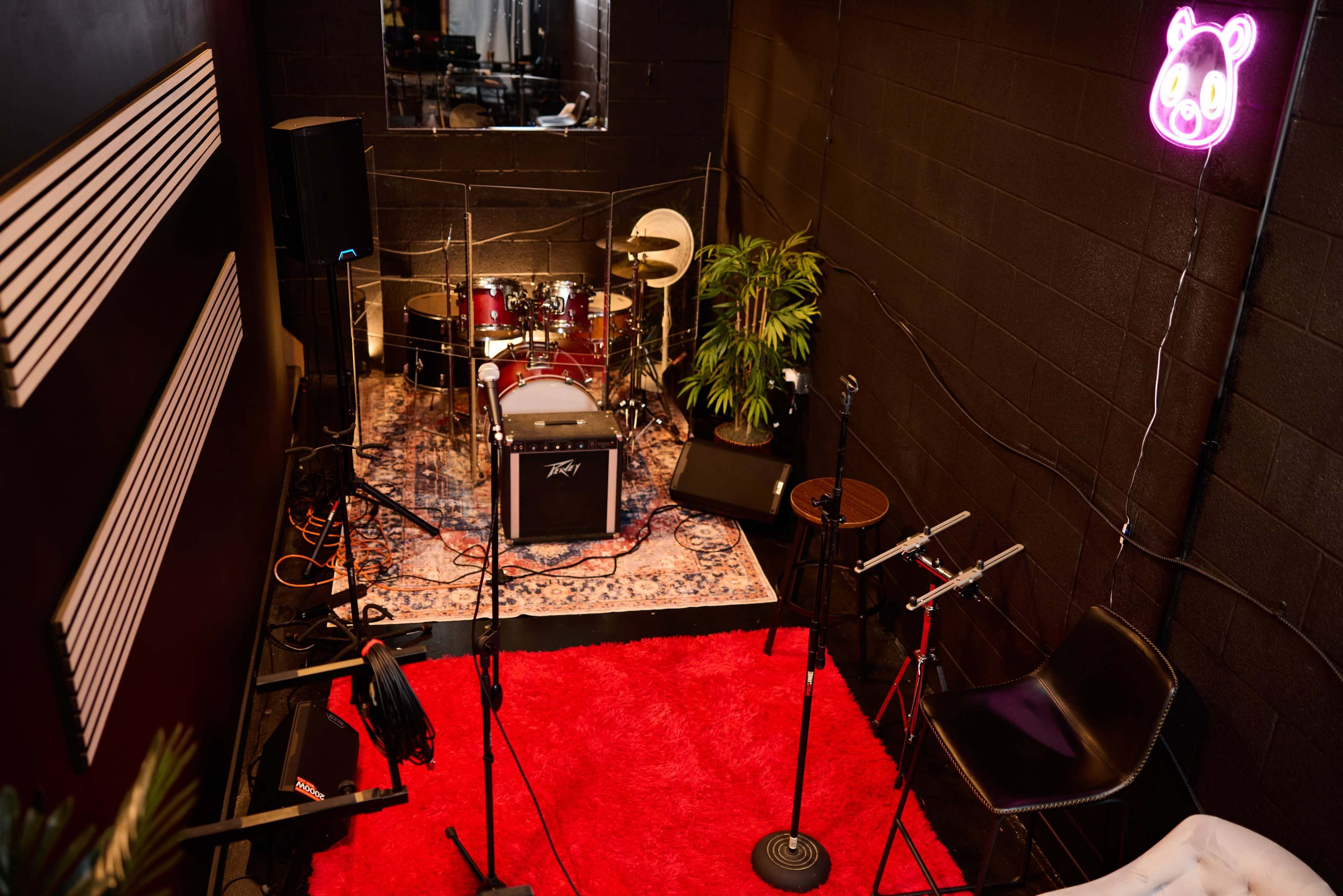 A small music rehearsal space features a drum set, microphones on stands, and a neon panda light against a black wall.
