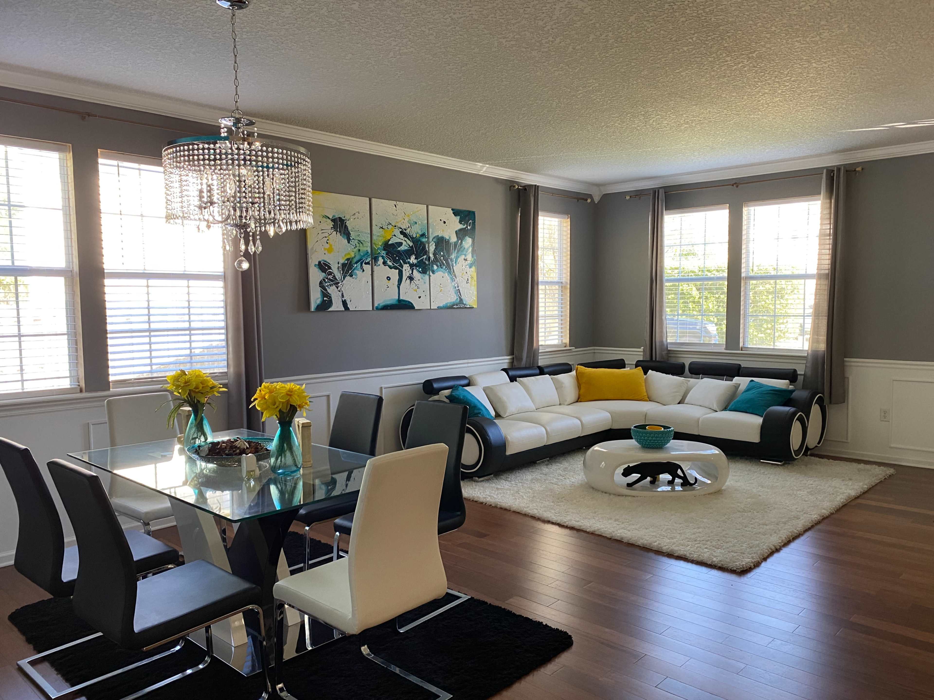 A modern living and dining area with a large sectional sofa, a dining table set for six, and a chandelier overhead, accented by a colorful wall art piece.