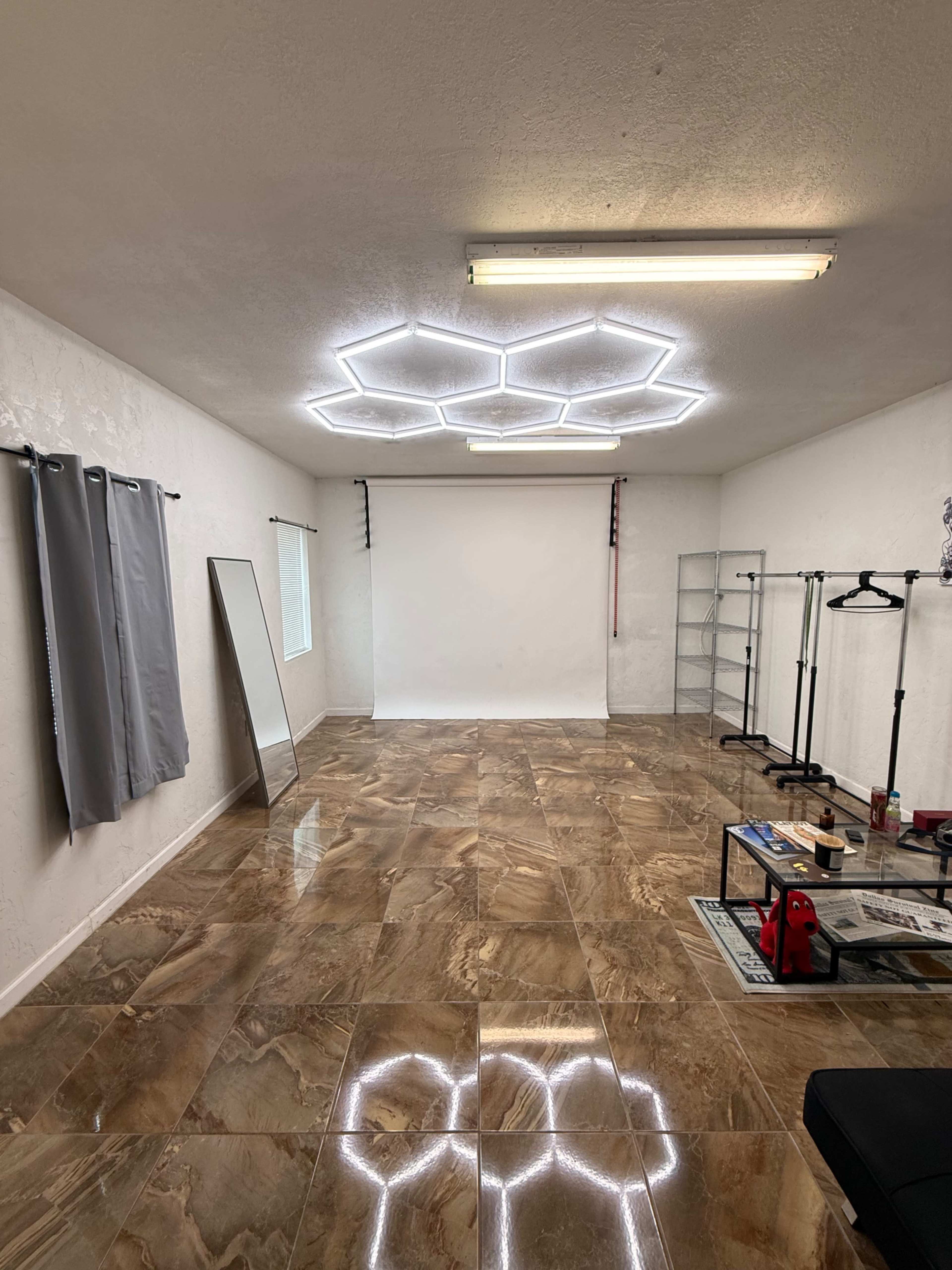 The image shows an empty room with glossy tiled floors, a light-colored wall, a hexagonal ceiling light design, a large mirror, and some clothing racks along with a backdrop setup.